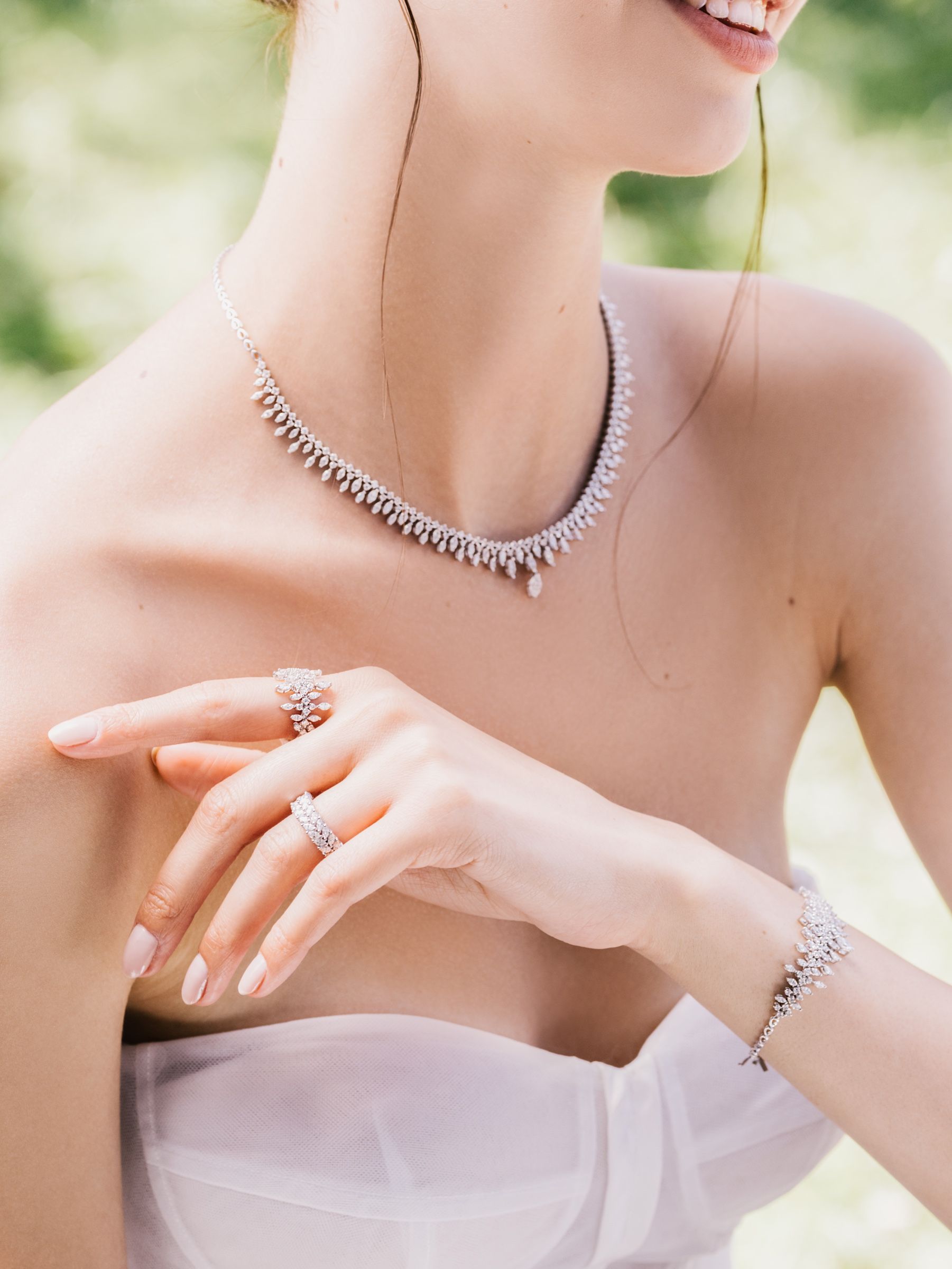 Close frame of the model's neck and hand: a diamond fringe necklace, two rings and a matching bracelet above a strapless white gown.