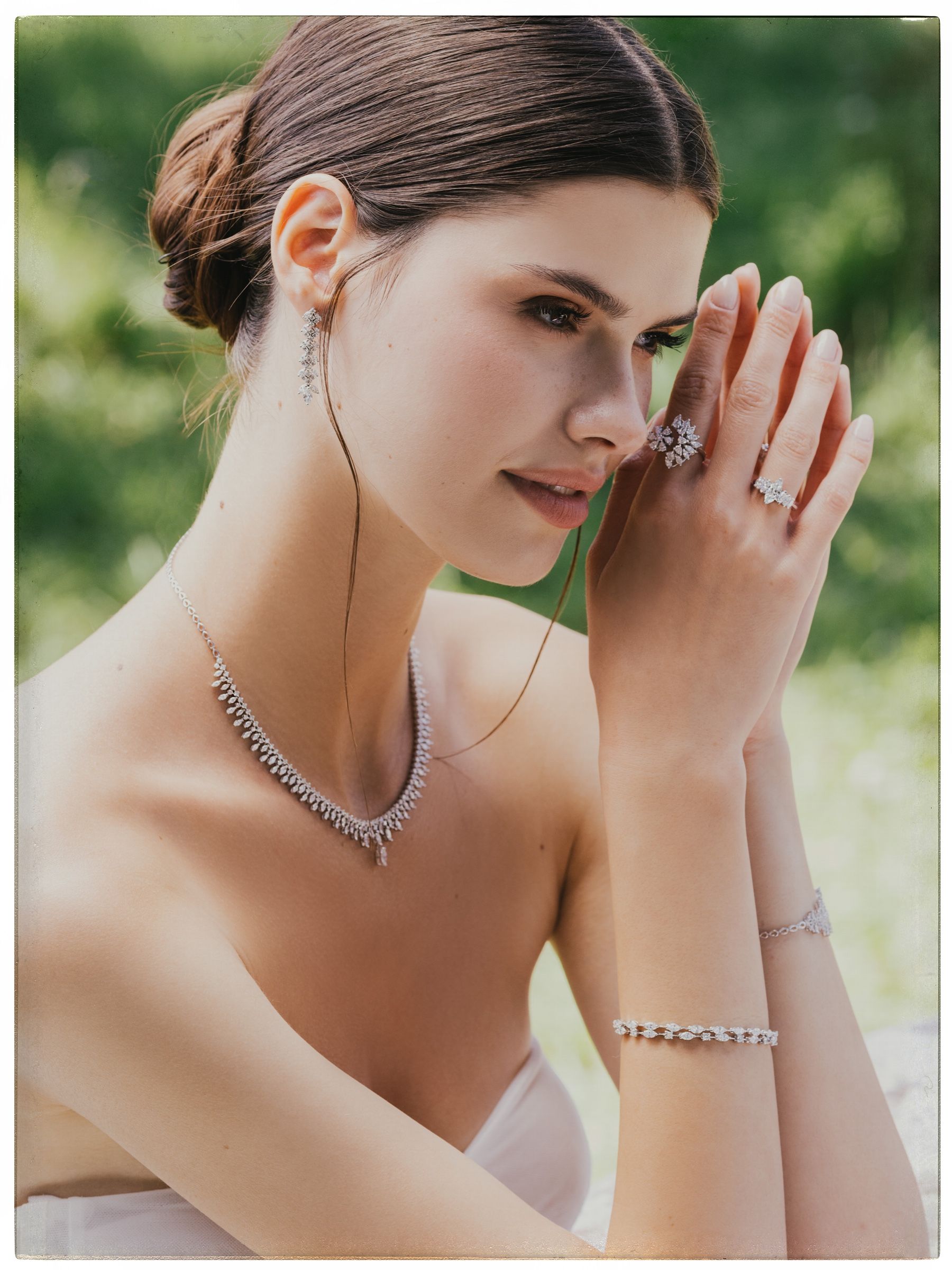 The model presses her hands together beside her face, wearing a diamond drop earring, fringe necklace, two rings and two bracelets above a strapless gown.