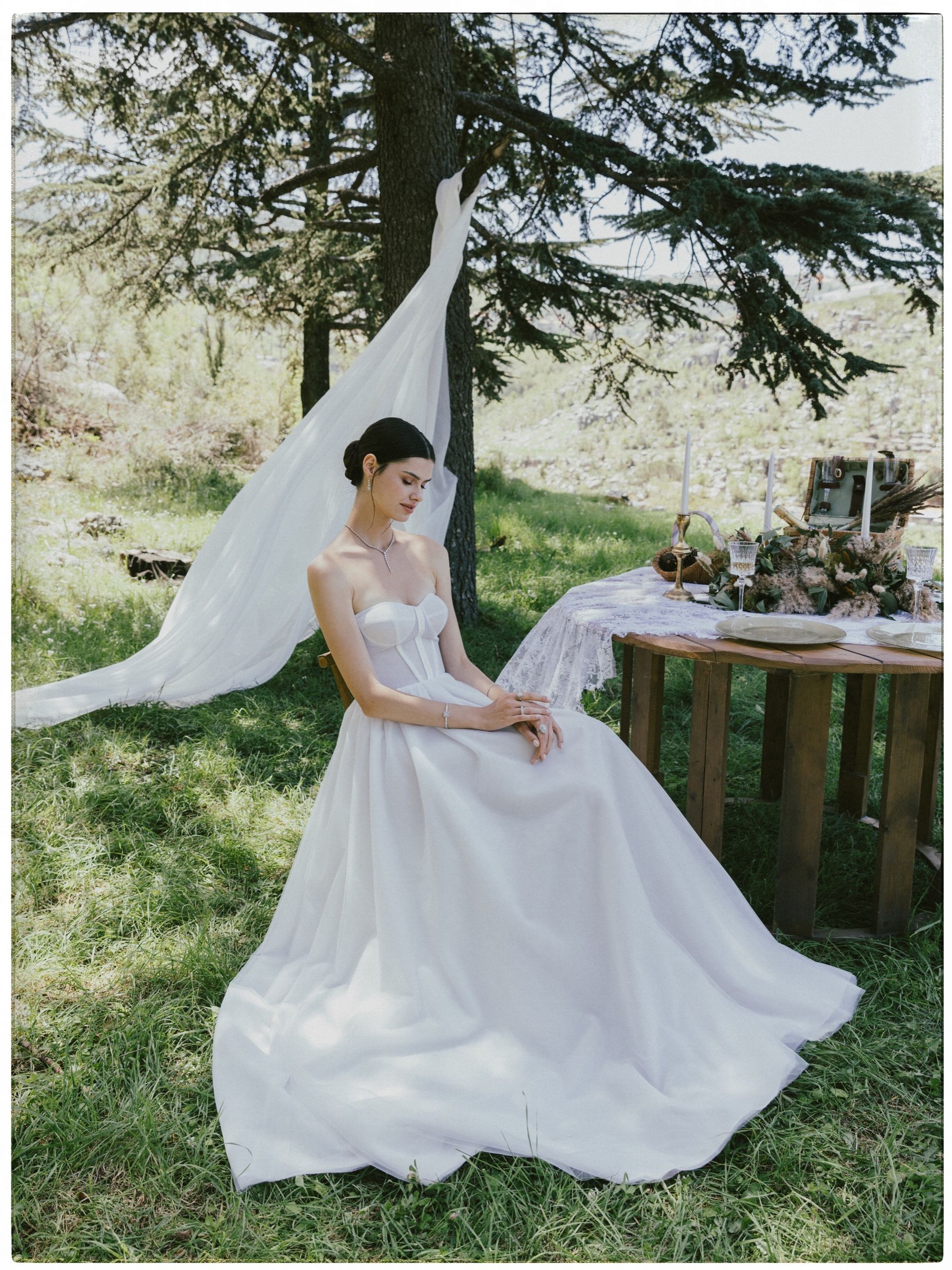 Seated beside a laid table in a full white gown, veil caught on a cedar branch, she wears a diamond drop necklace, bracelet and rings.