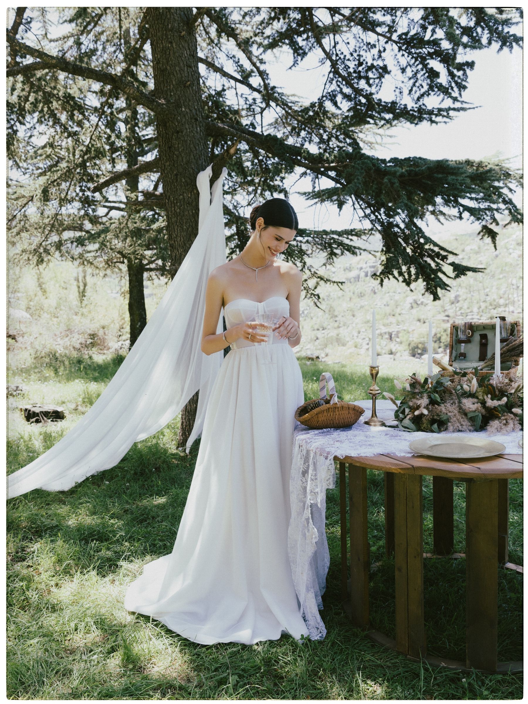 Under a cedar, veil hung from the trunk, she holds a coupe glass in a strapless white gown, wearing a diamond drop necklace and bracelet.