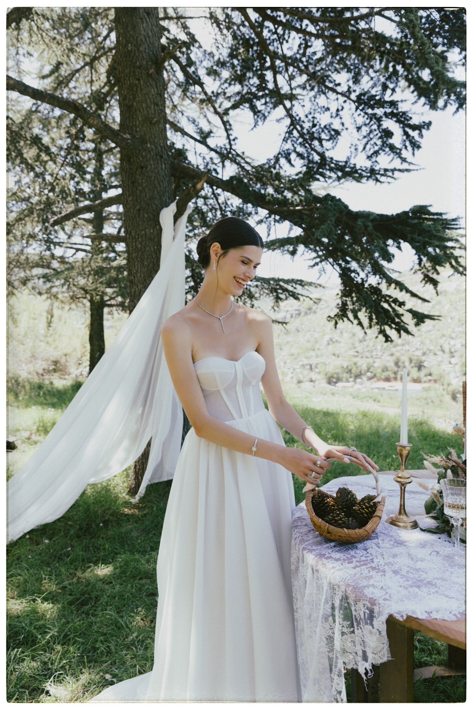 Laughing, the model reaches for a basket of pine cones on a lace table, wearing a diamond drop necklace, bracelets and rings on both hands.