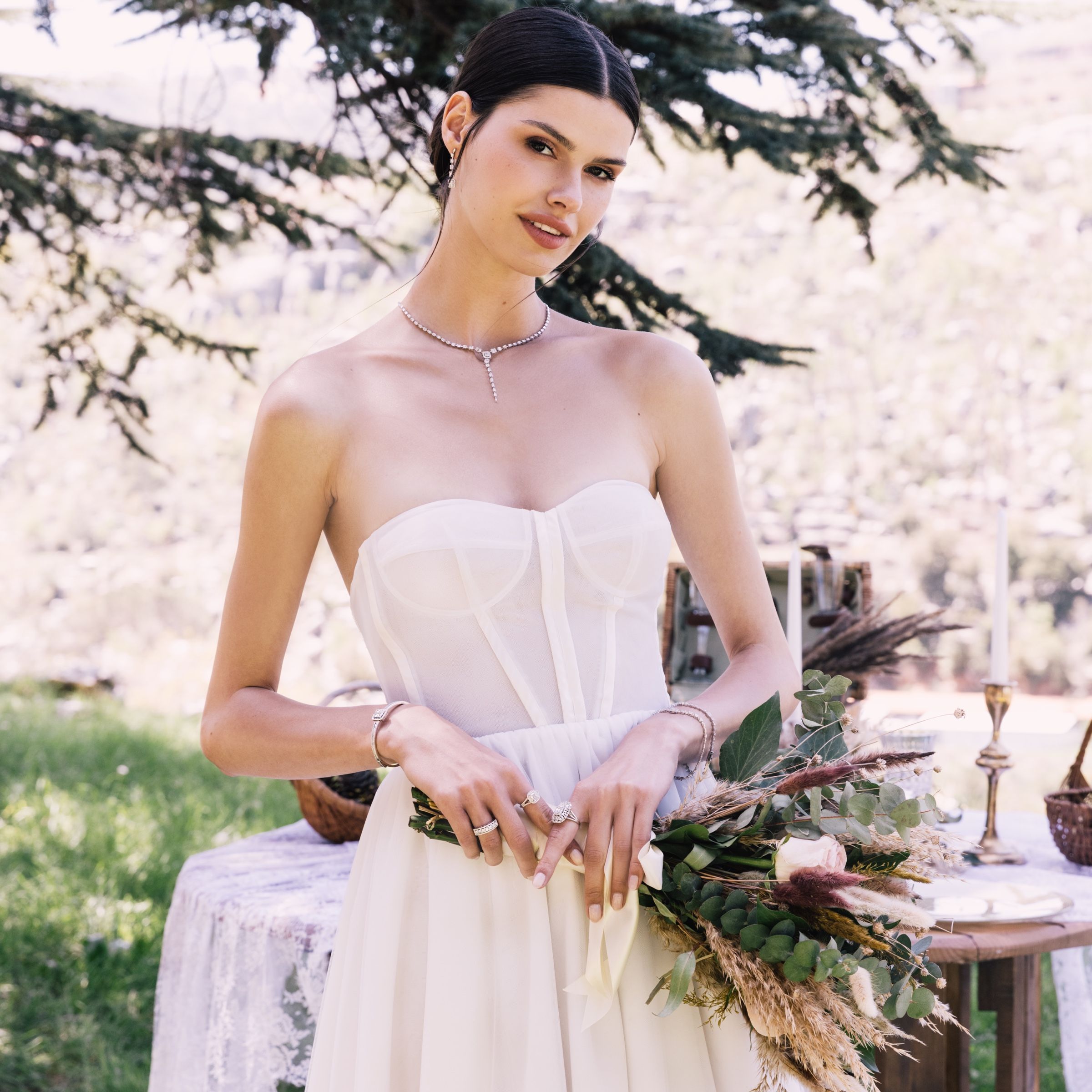 Beneath a cedar branch, she holds a large eucalyptus bouquet in a corseted white gown, wearing a diamond drop necklace, bracelets and several rings.