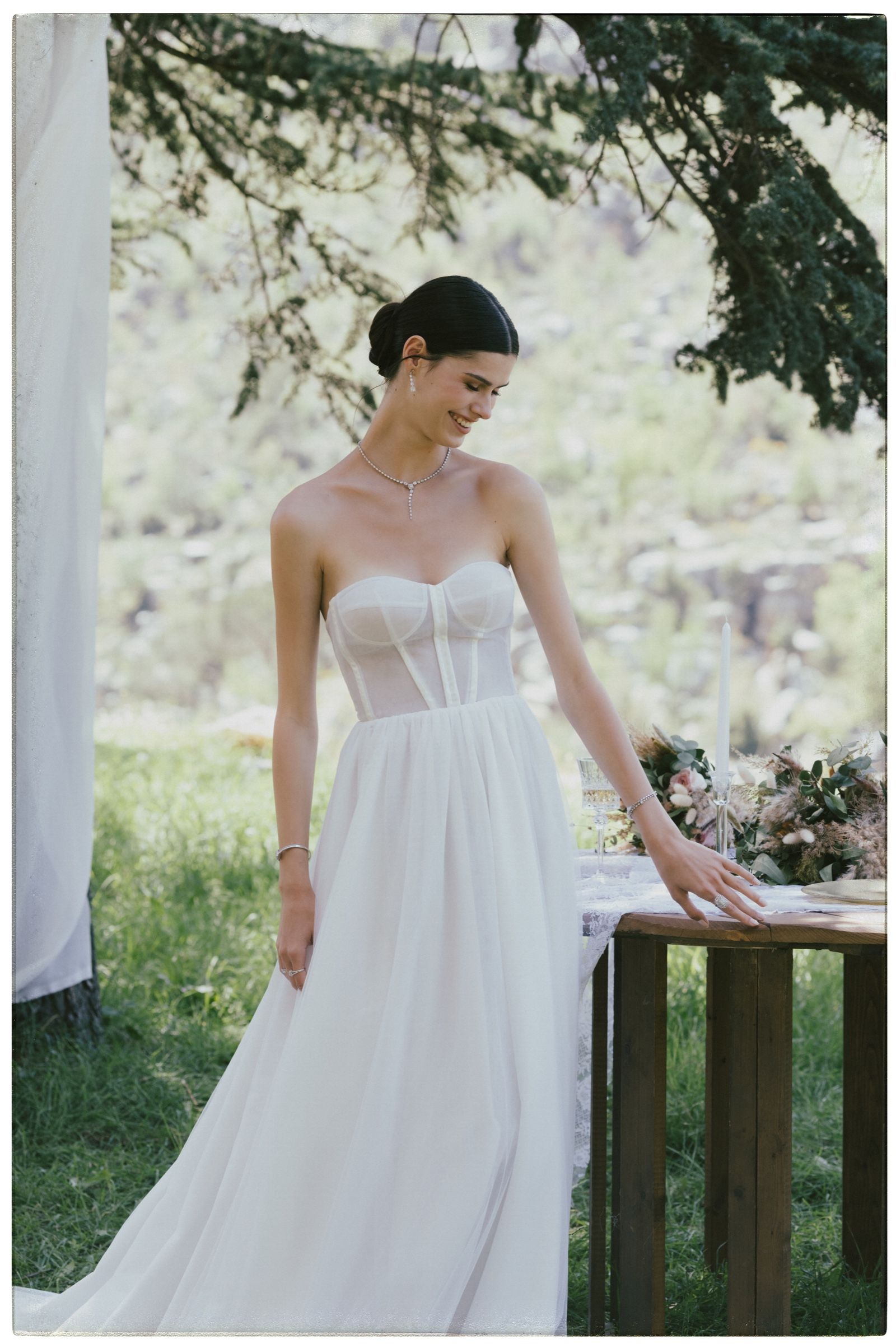 The model rests a ringed hand on the laid table in a strapless white gown, wearing a diamond drop necklace and slim bracelets on both wrists.