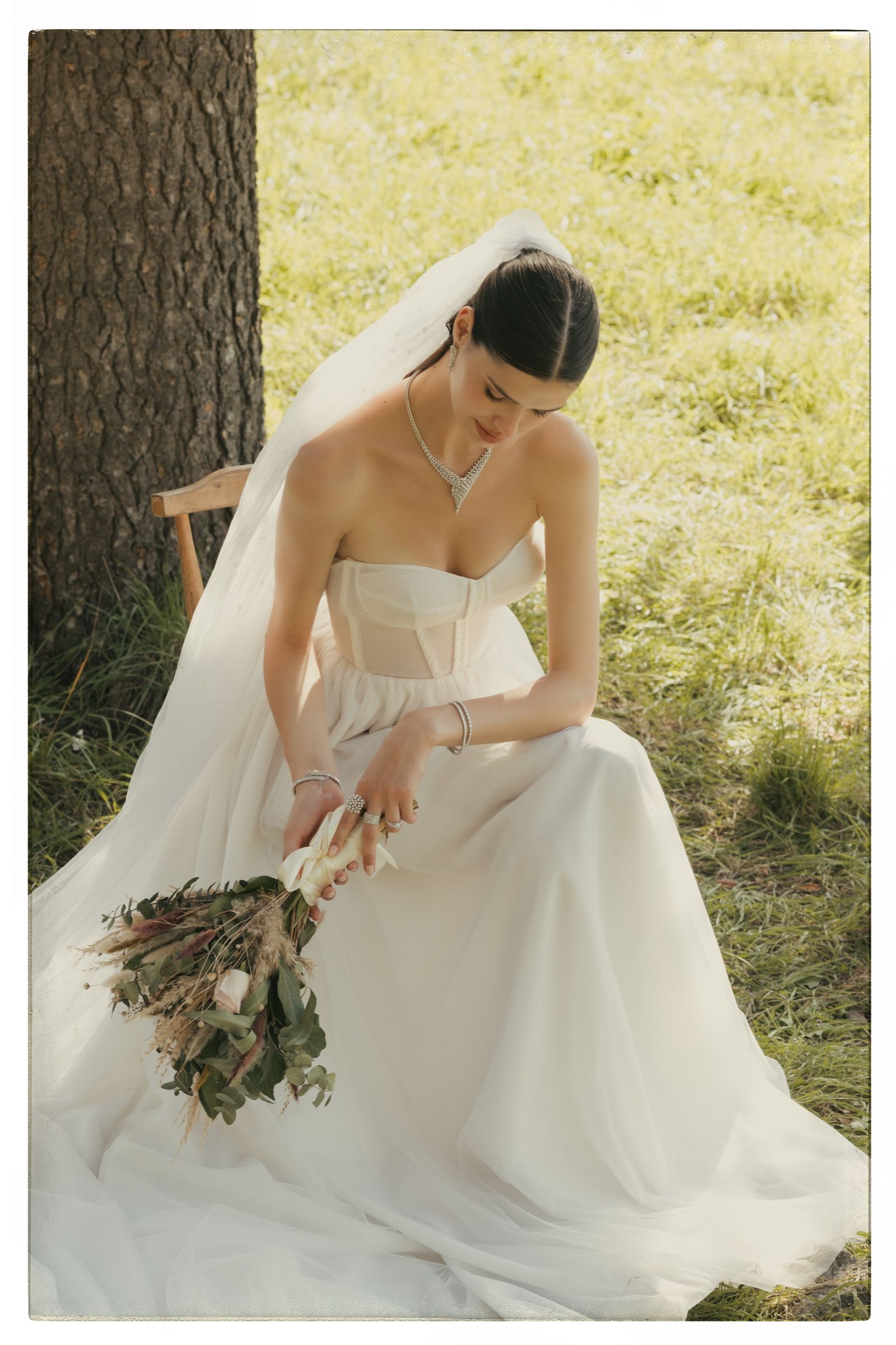 The model sits on a wooden chair in the grass, veil trailing, lowering a bouquet and wearing a V-shaped diamond necklace, bracelets and rings.