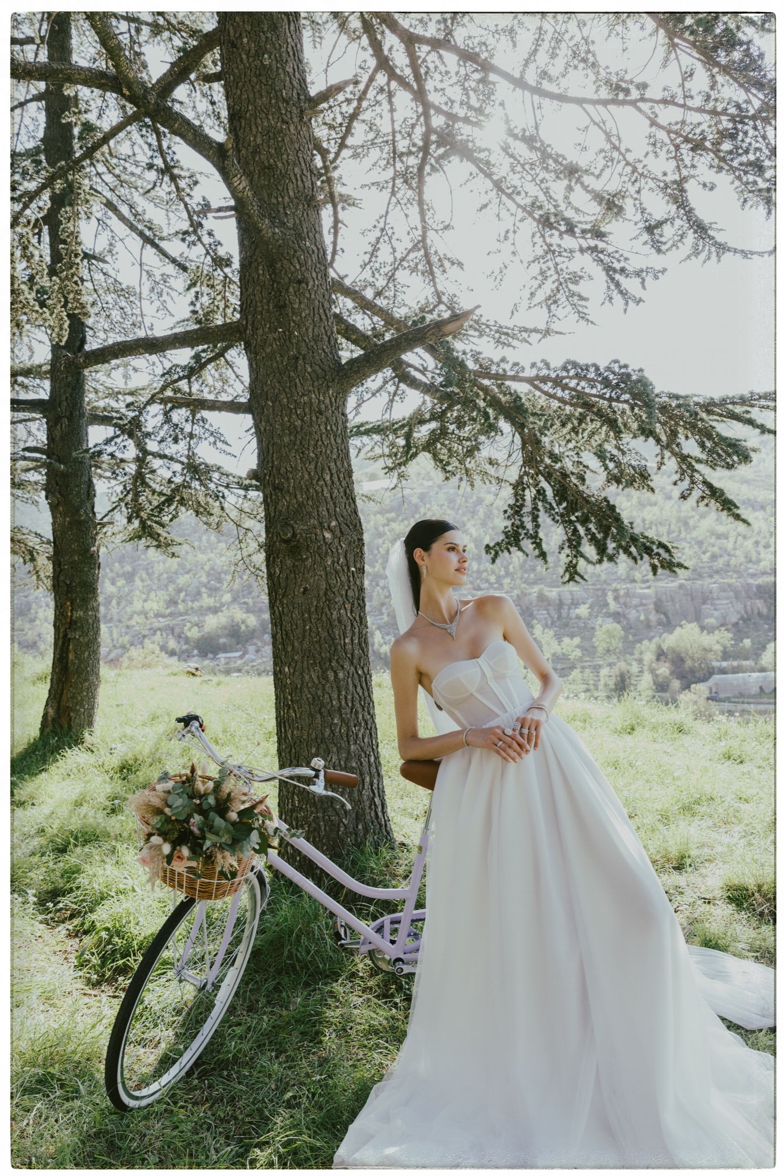 In a strapless tulle gown and veil, the model stands beside a lilac bicycle with a flower-filled basket beneath cedars, wearing a diamond necklace and rings.