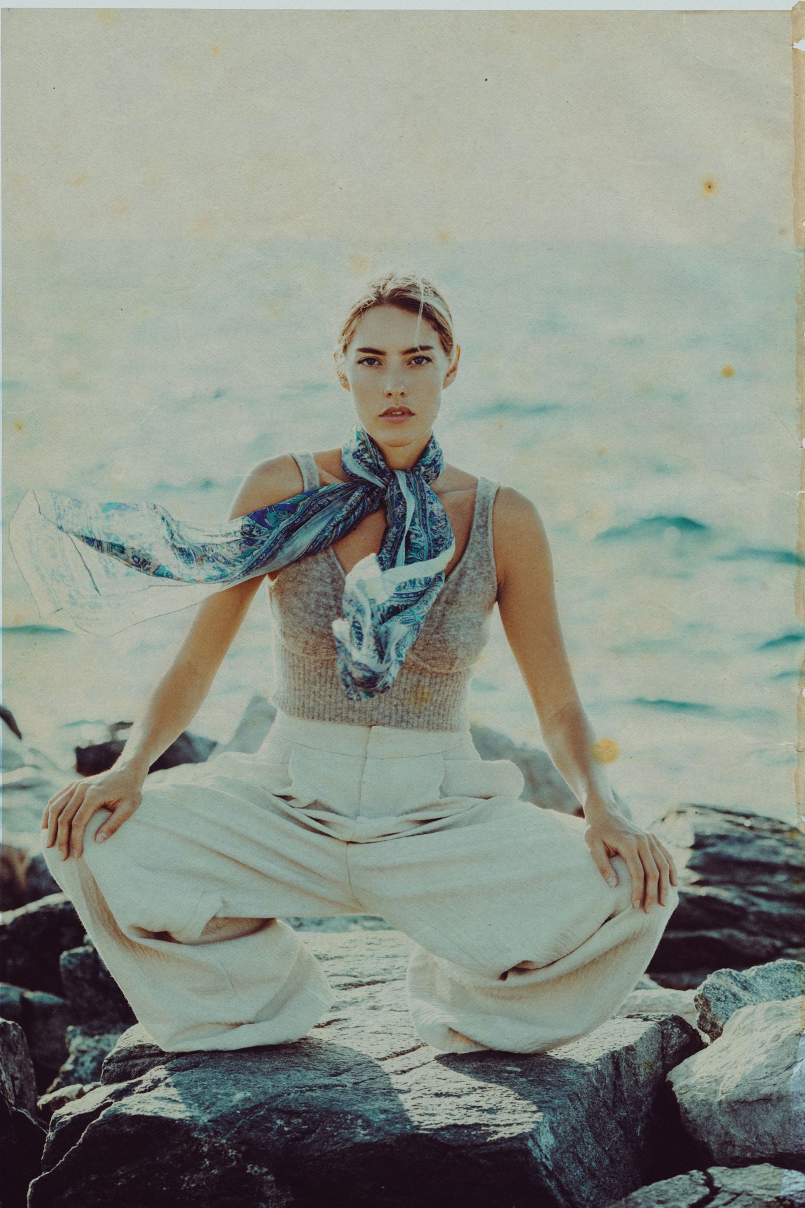 Gio sits cross-legged on rocks above the sea, a blue paisley scarf flying out from her neck over a grey knit vest and wide cream trousers.