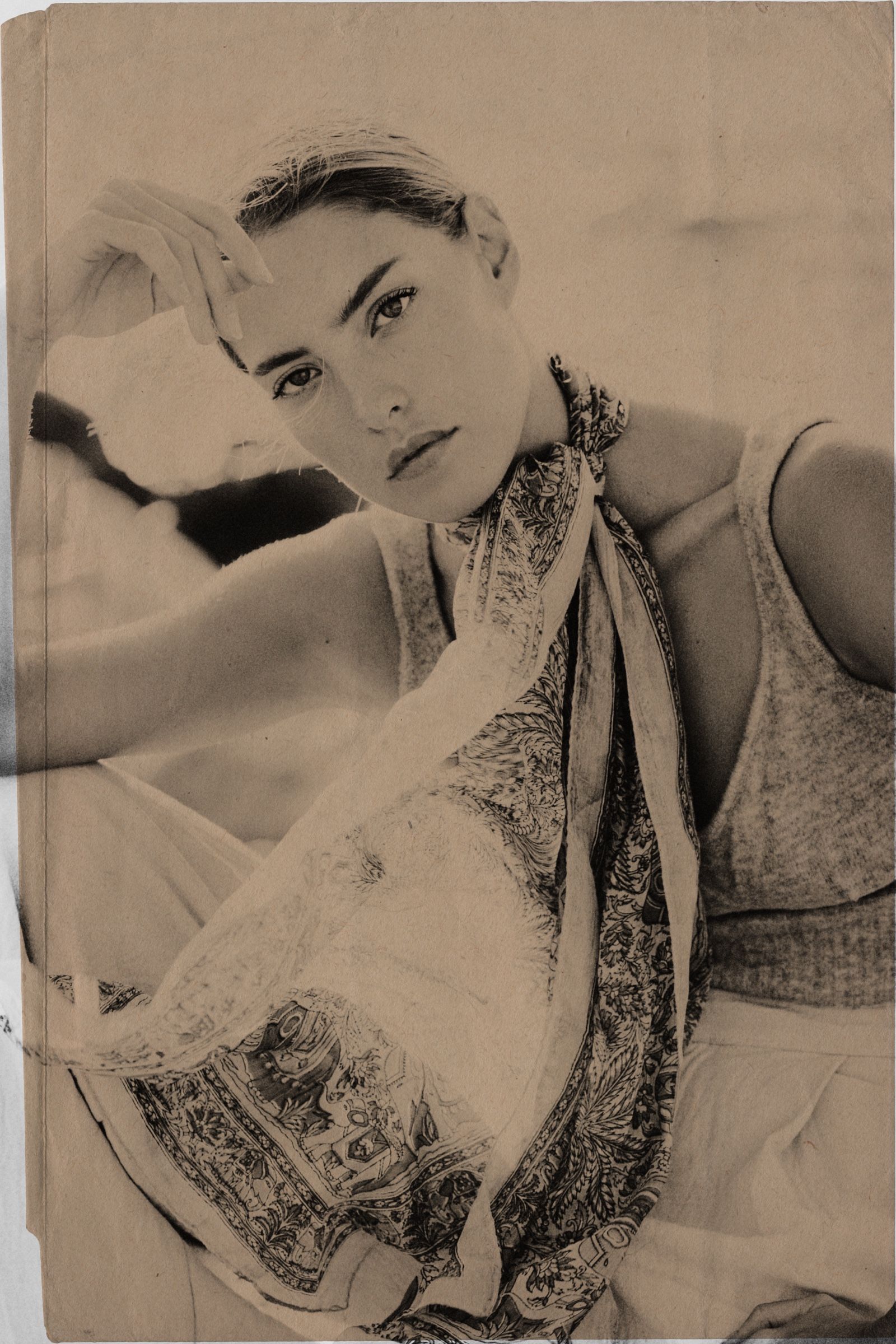 Sepia, printed on folded and foxed paper — a hand shading her brow, the scarf falling from one shoulder, looking straight out.