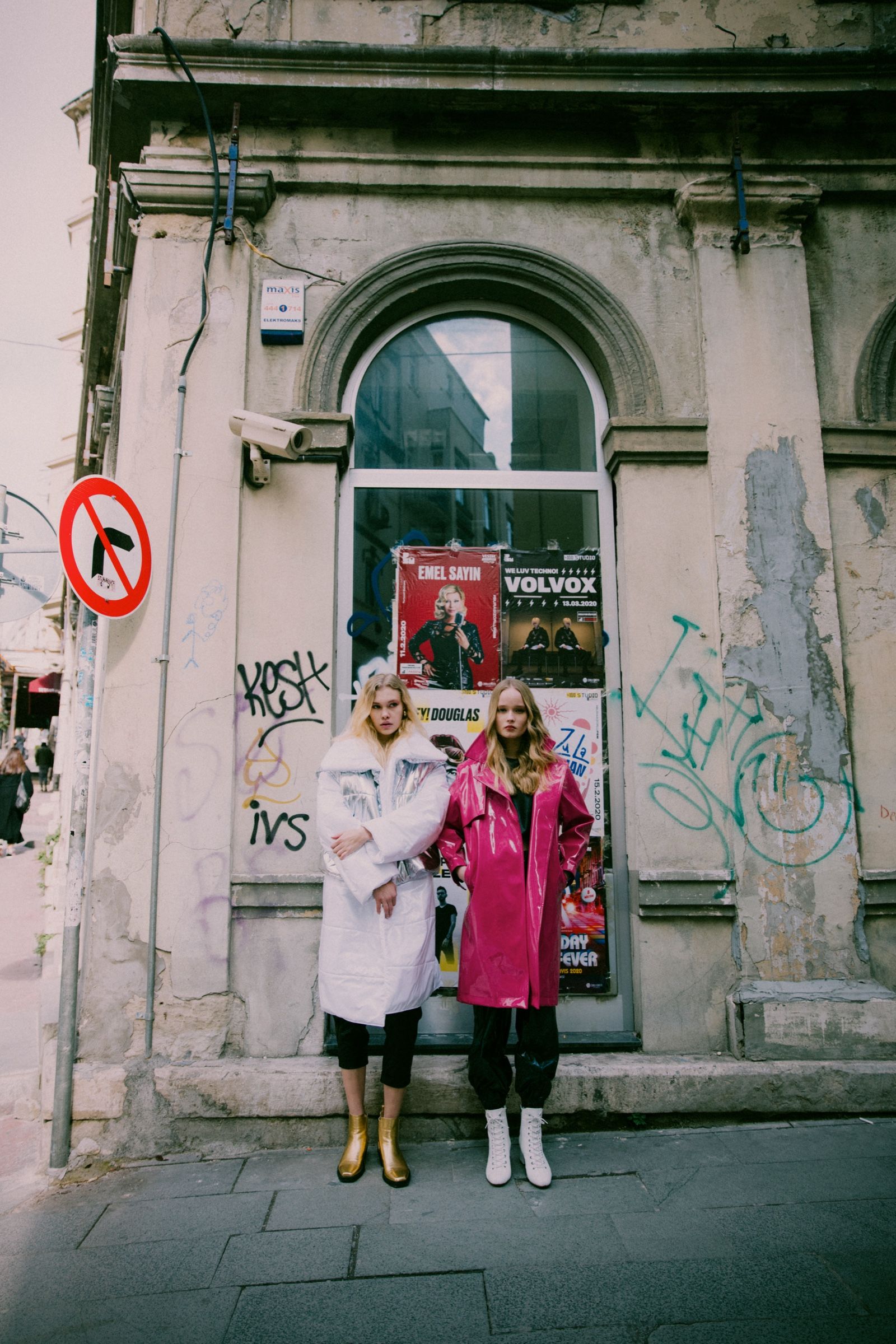 Two models, one in a white and silver puffer coat, one in a hot pink vinyl trench, stand before a graffitied storefront pasted with concert posters.