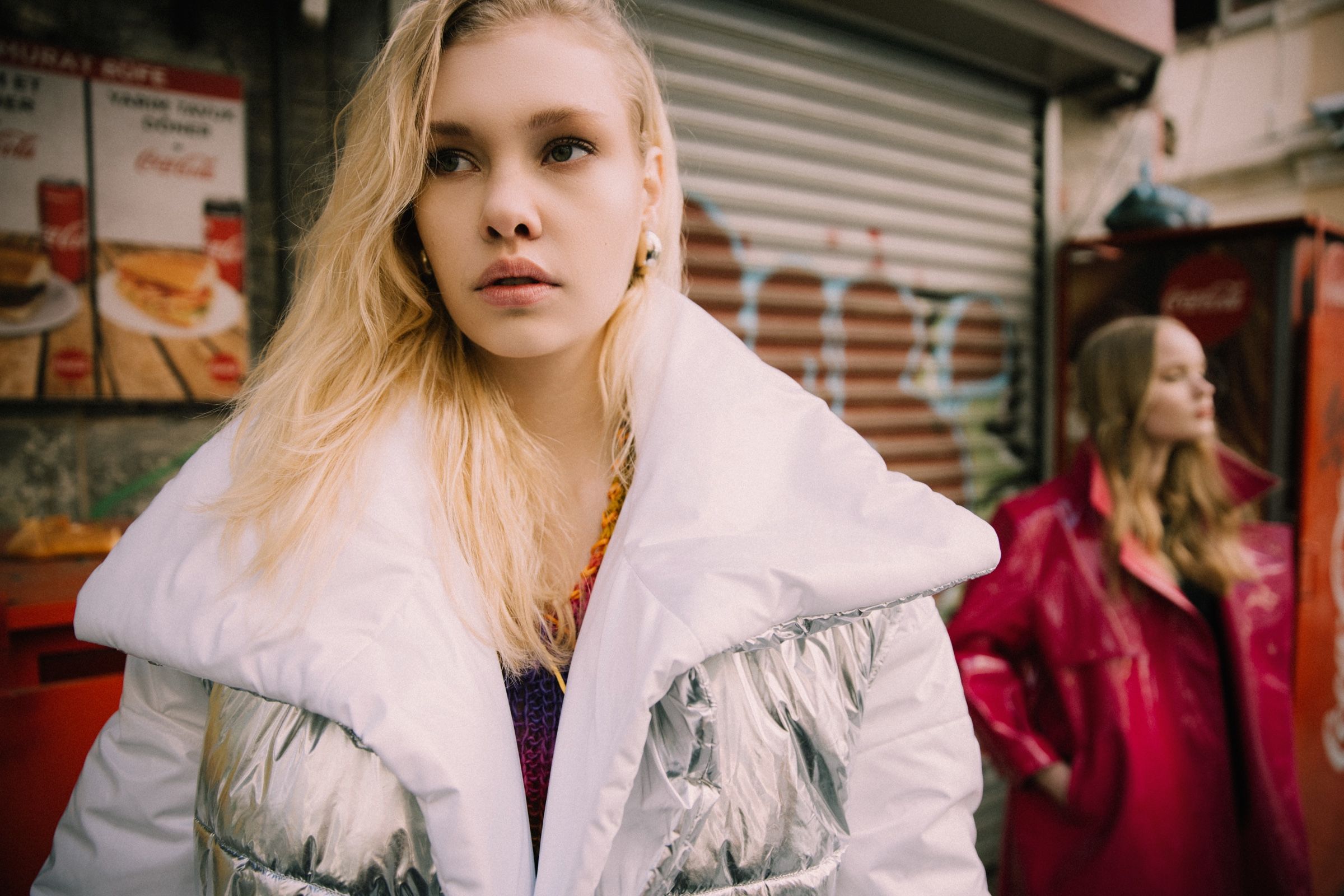 The white puffer's oversized collar and silver quilting fill the frame; the second model, in the pink vinyl trench, stands blurred by a shuttered cafe.