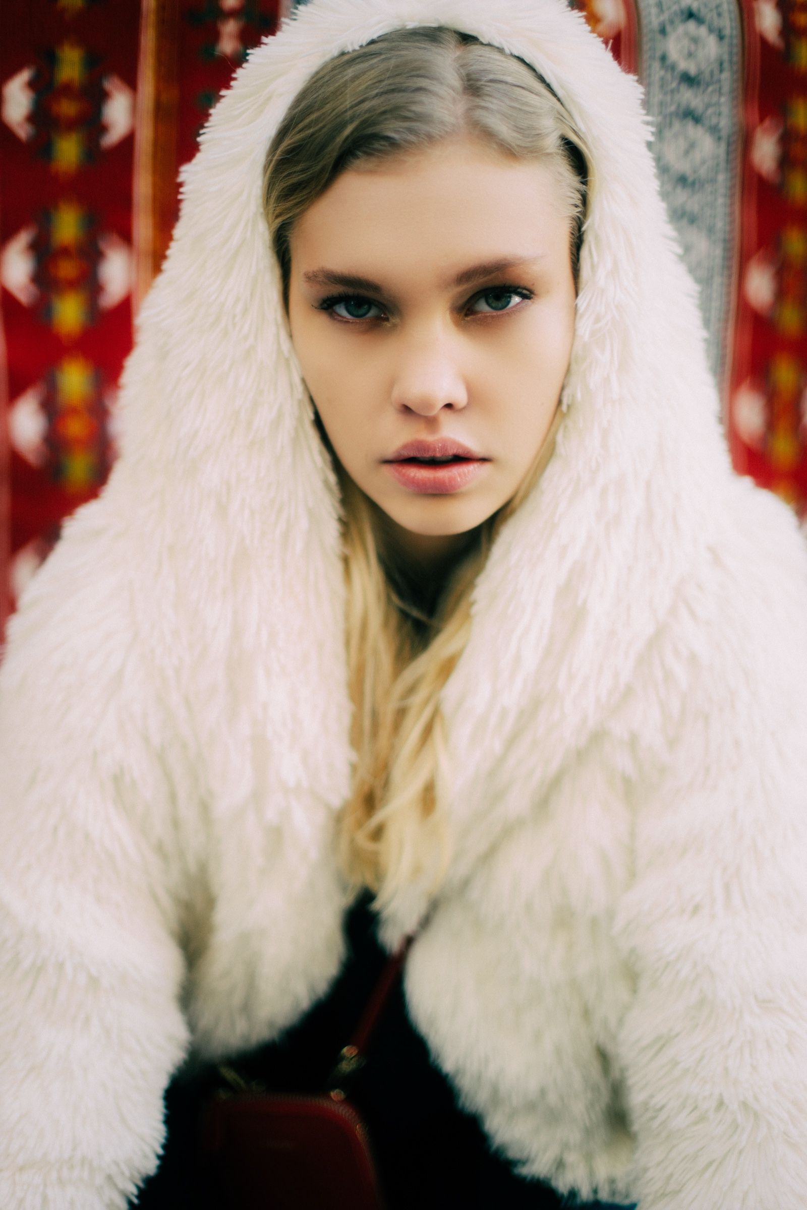 A white shaggy fur coat with its wide collar pulled up over the model's head, framing her face against hanging red patterned carpets.