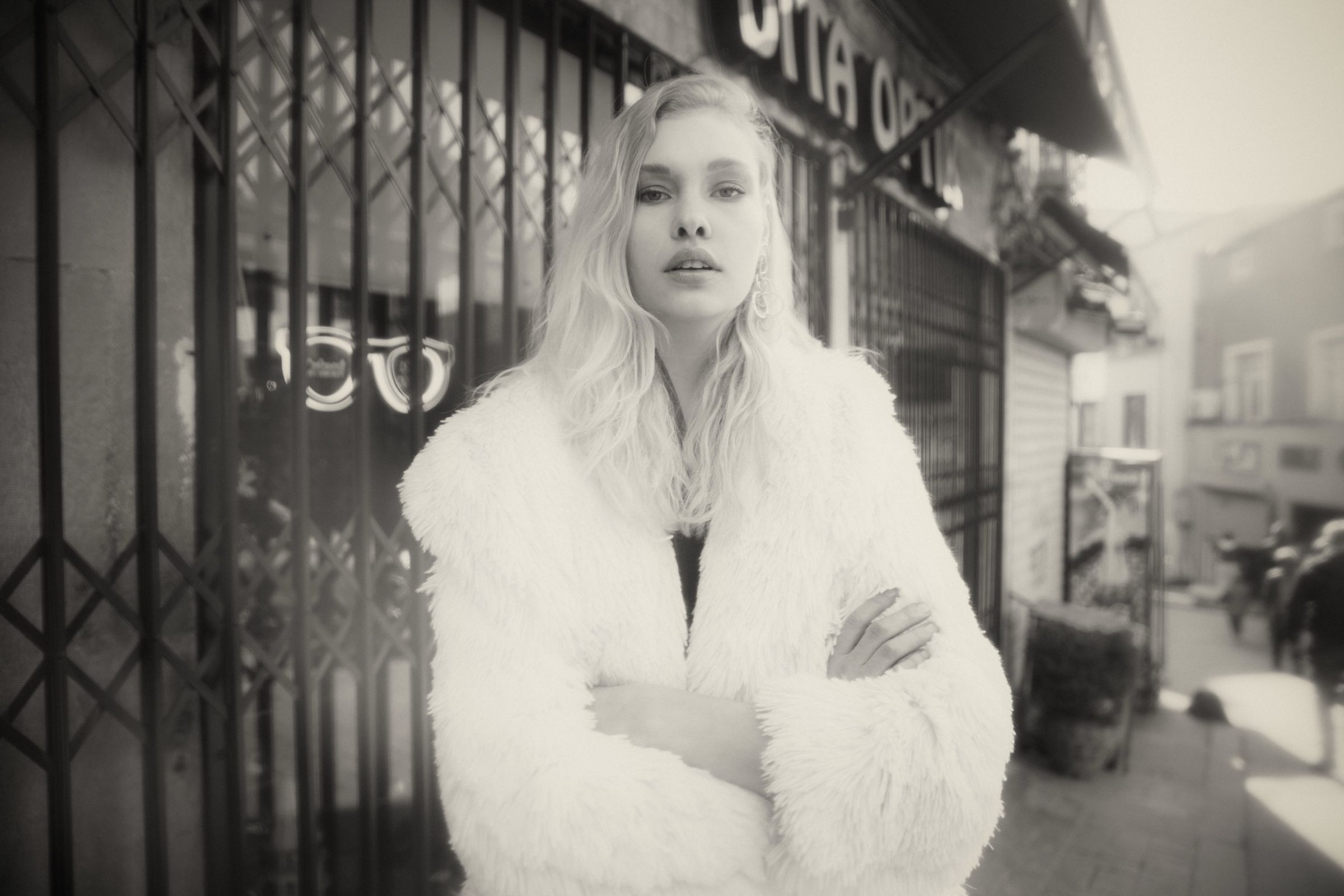 Black and white: a model in a long white shaggy fur coat stands arms folded outside a shuttered shopfront with spectacles painted on the glass.