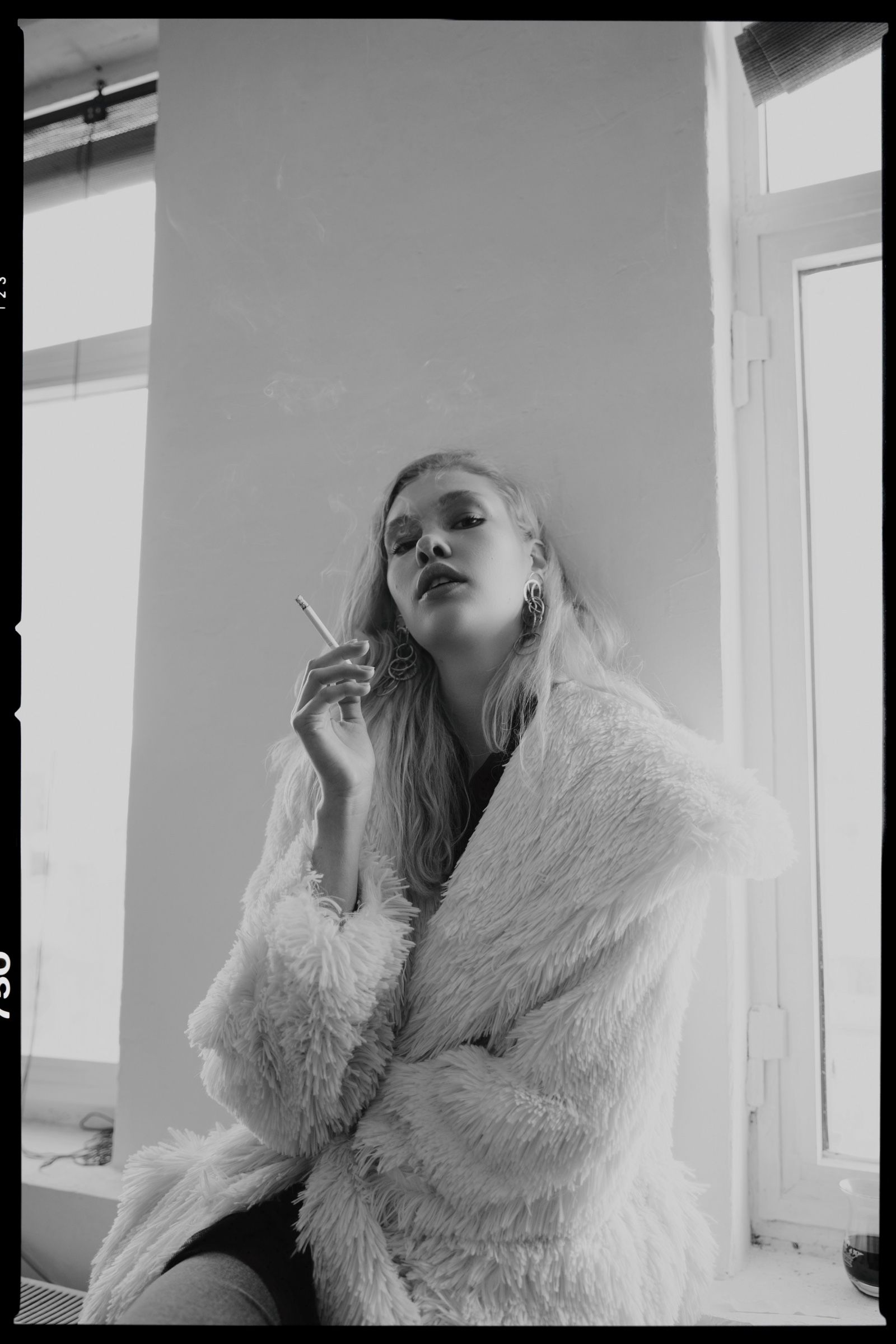 Black and white with the film edge showing: a model in the white shaggy fur coat smokes, seated against a wall beside a bright window.