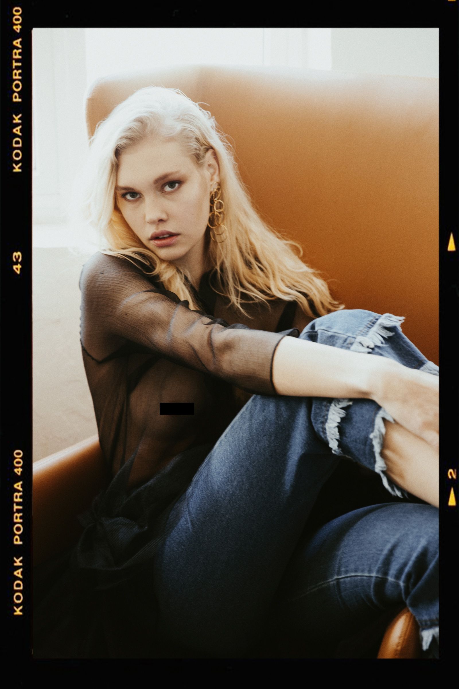 A sheer black mesh top and ripped blue jeans, the model curled on a tan leather sofa, a Kodak Portra 400 rebate down the edge.