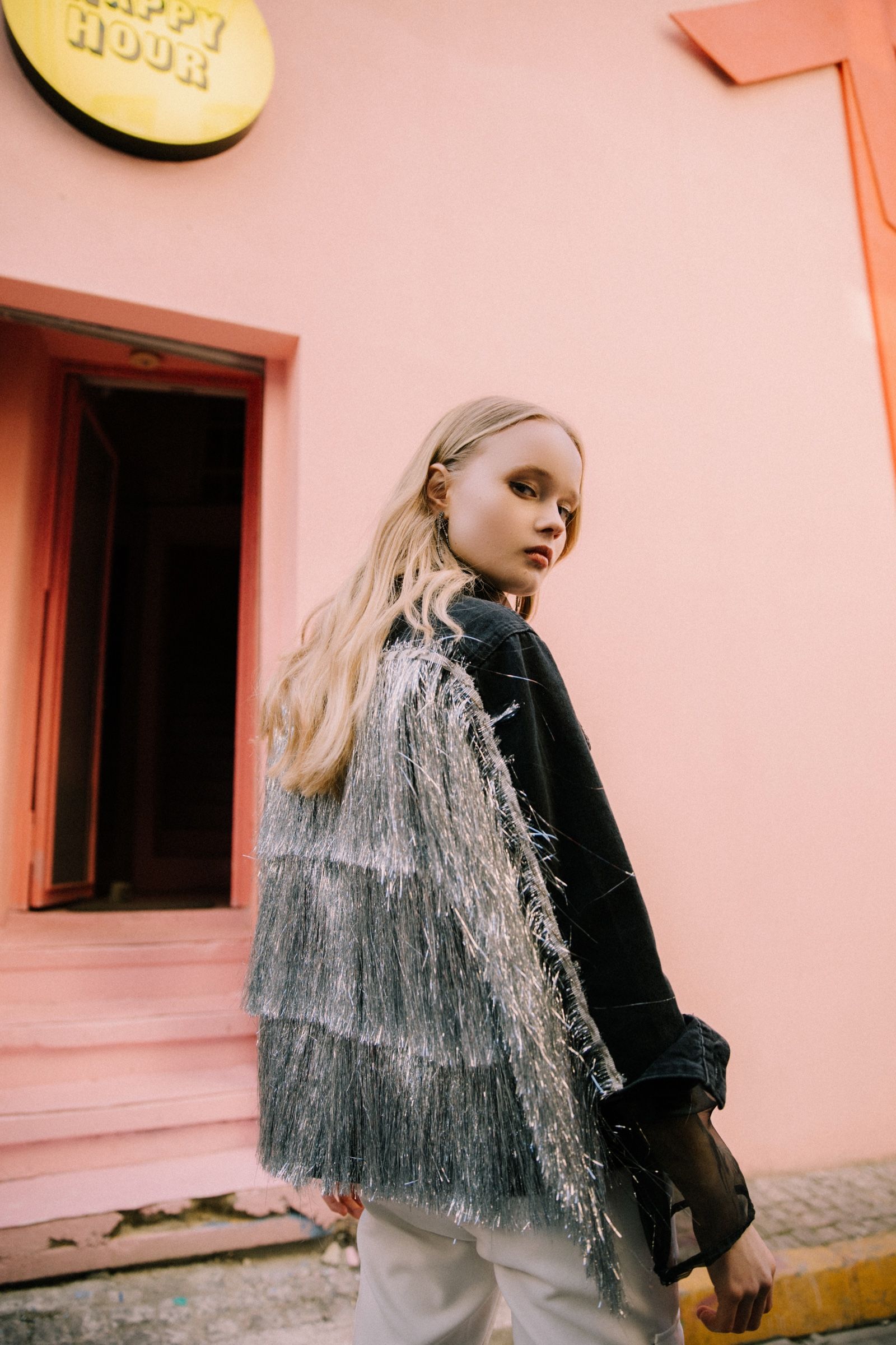 A black denim jacket with tiered silver tinsel fringe and white trousers, the model turning by a pink doorway under a yellow Happy Hour sign.
