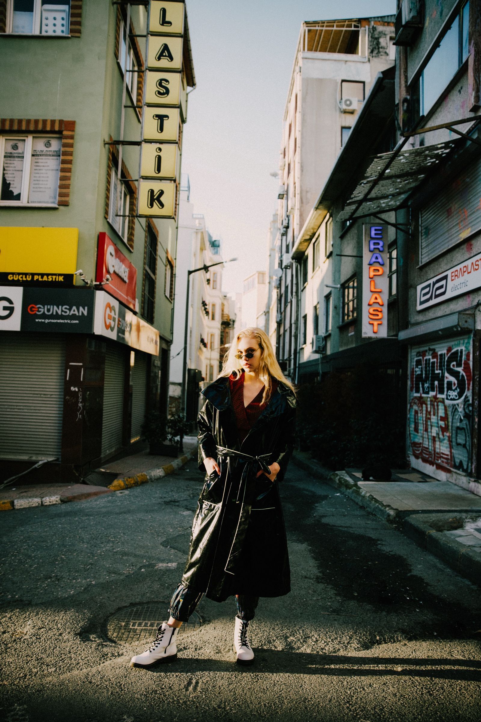 A belted black vinyl trench, red top and white lace-up boots, the model standing mid-street between graffitied shutters under a vertical Lastik sign.