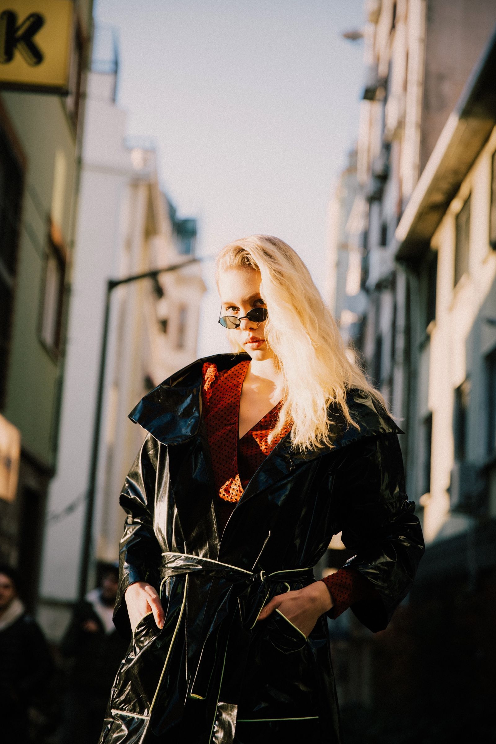 Closer: the black patent trench belted over a red polka-dot top, oversized collar upturned, hands in the pockets, narrow oval sunglasses, on a sunlit street.