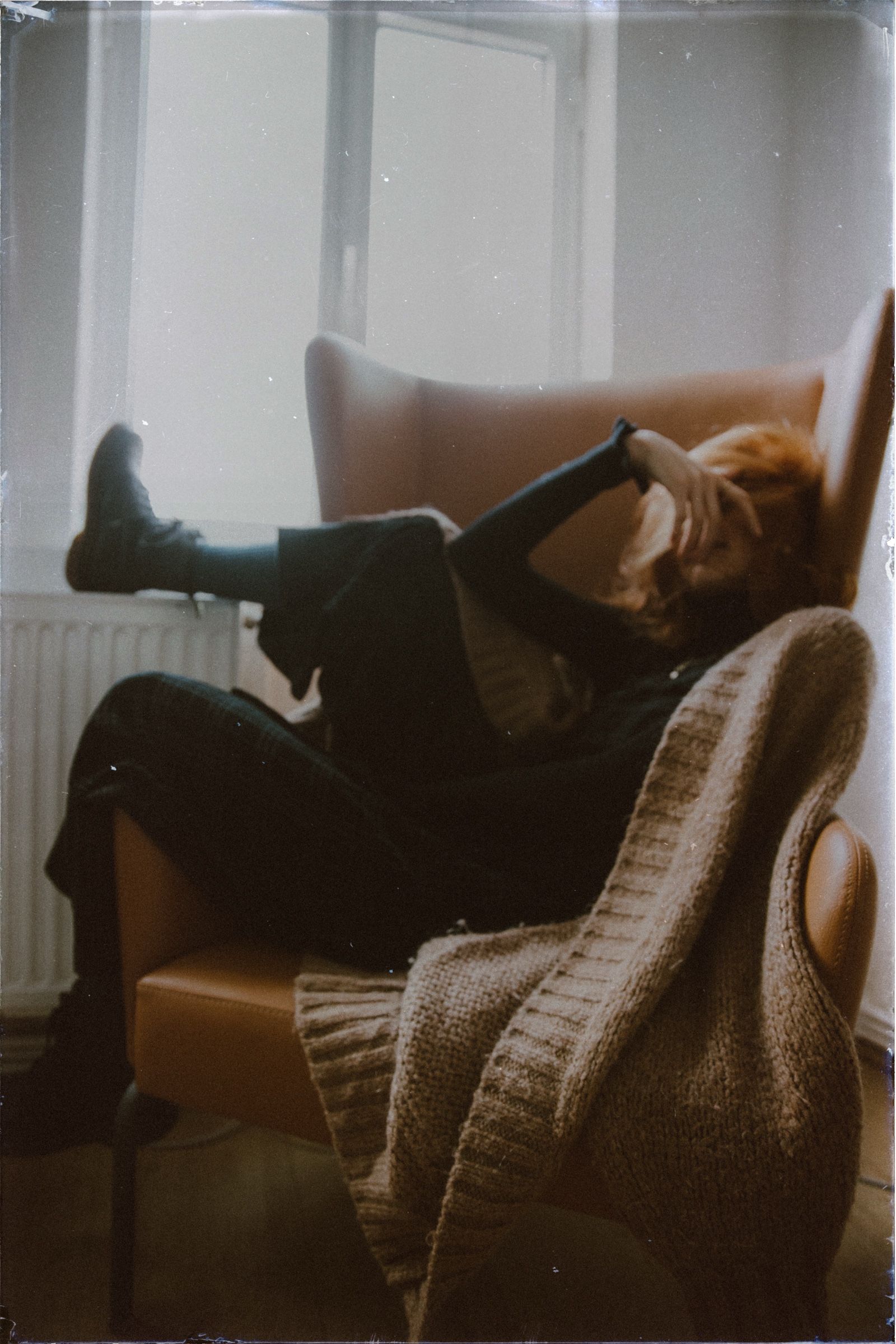 Elif lies back in a tan leather armchair, boots up on the radiator, one arm covering her face against the bright window.