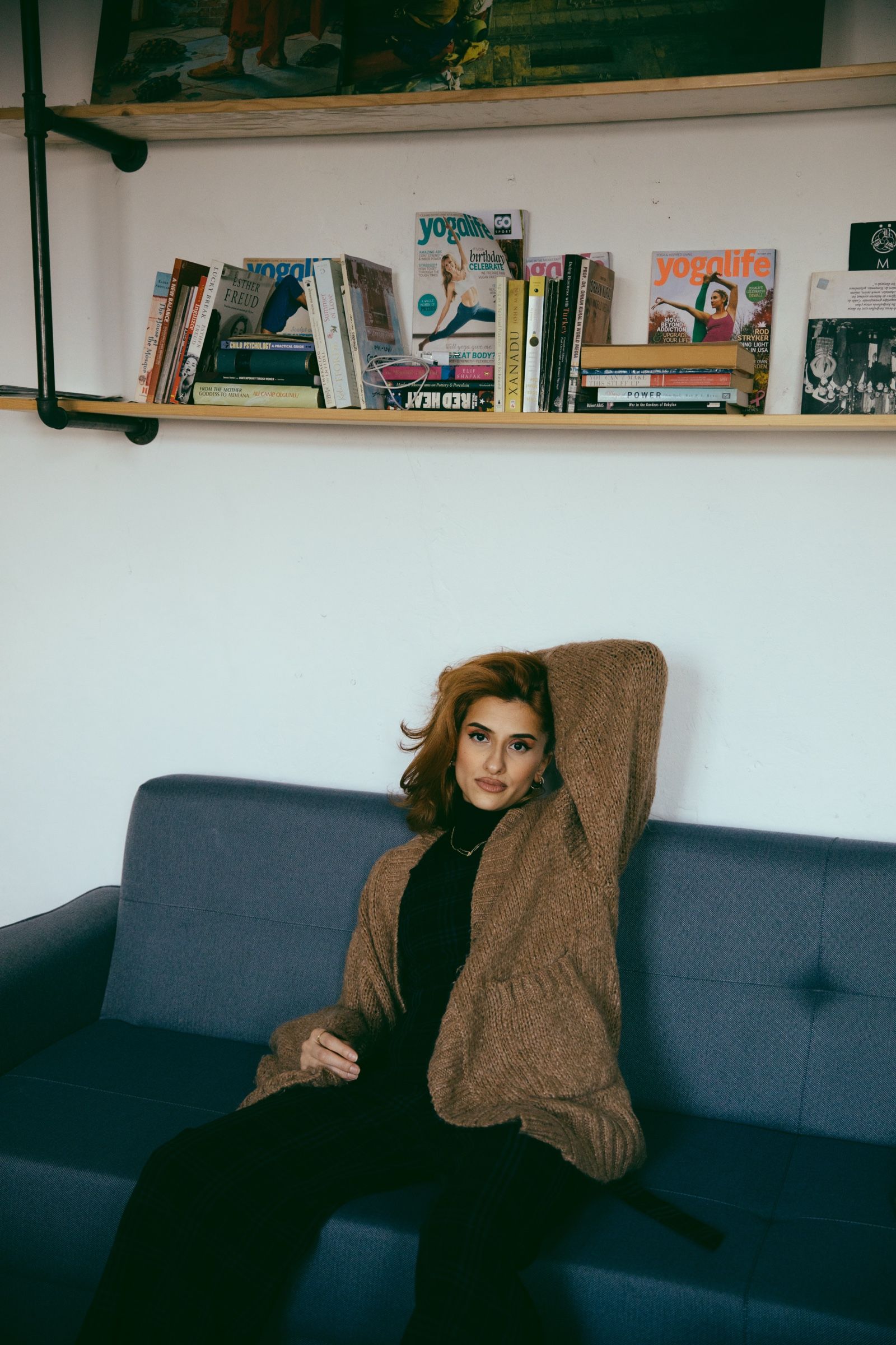 Elif sits on a blue sofa in a brown knit cardigan, one arm raised behind her head, shelves of books above.