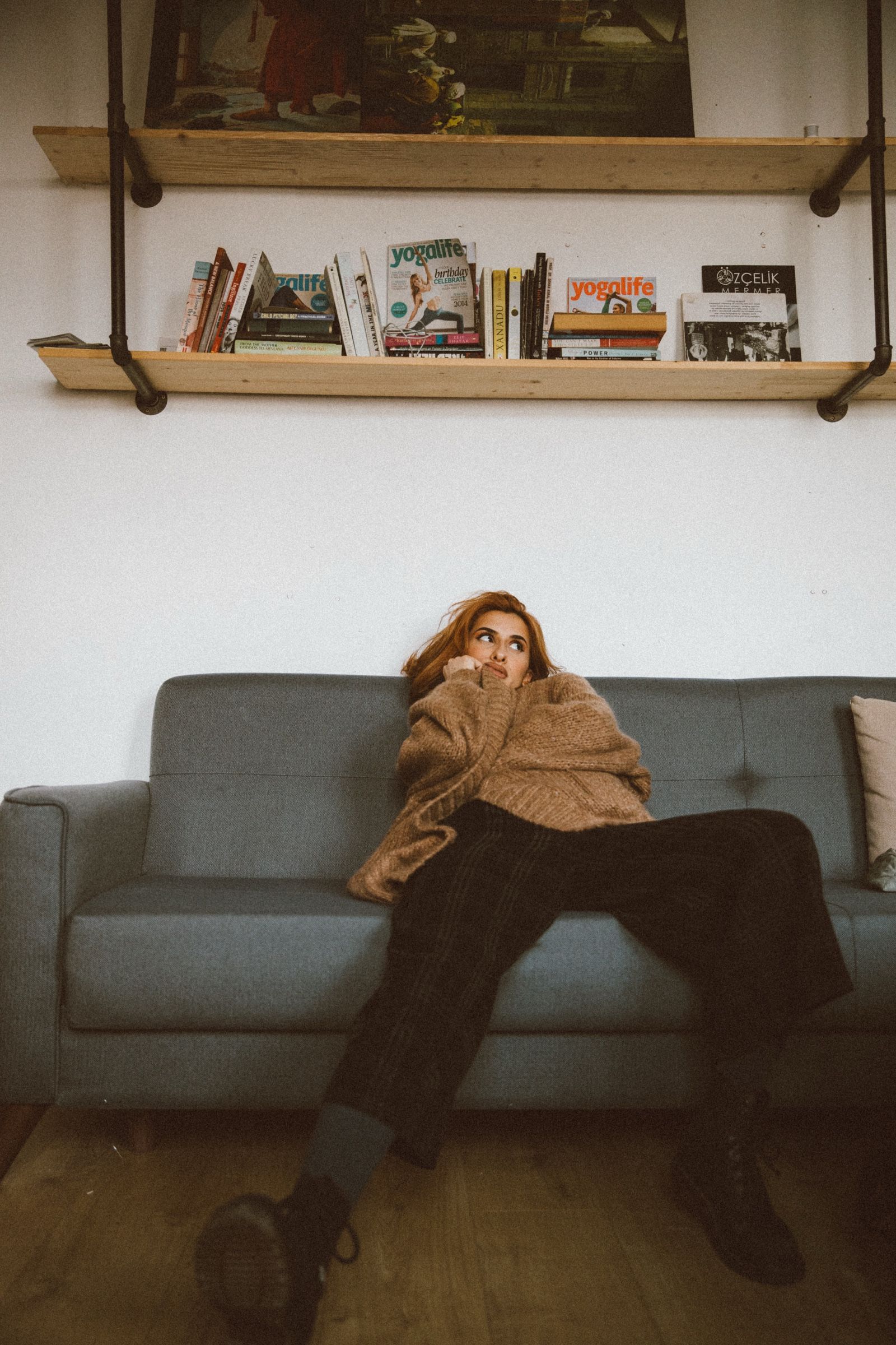 Elif sits low on a grey sofa in an oversized knit sweater, legs stretched out in boots, a shelf of books above.