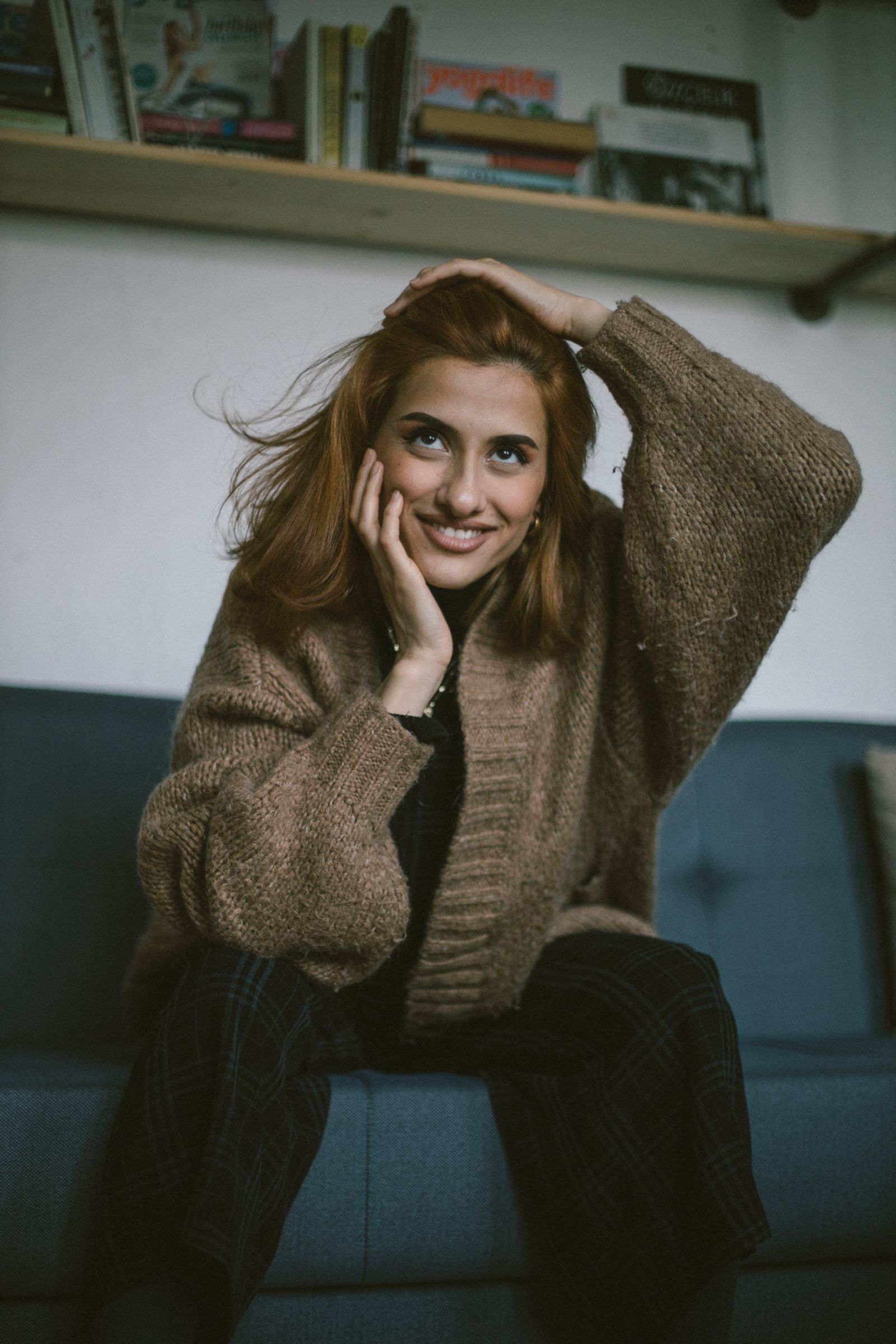 Elif smiles and looks up past the camera, one hand in her hair and the other at her cheek, on the blue sofa.