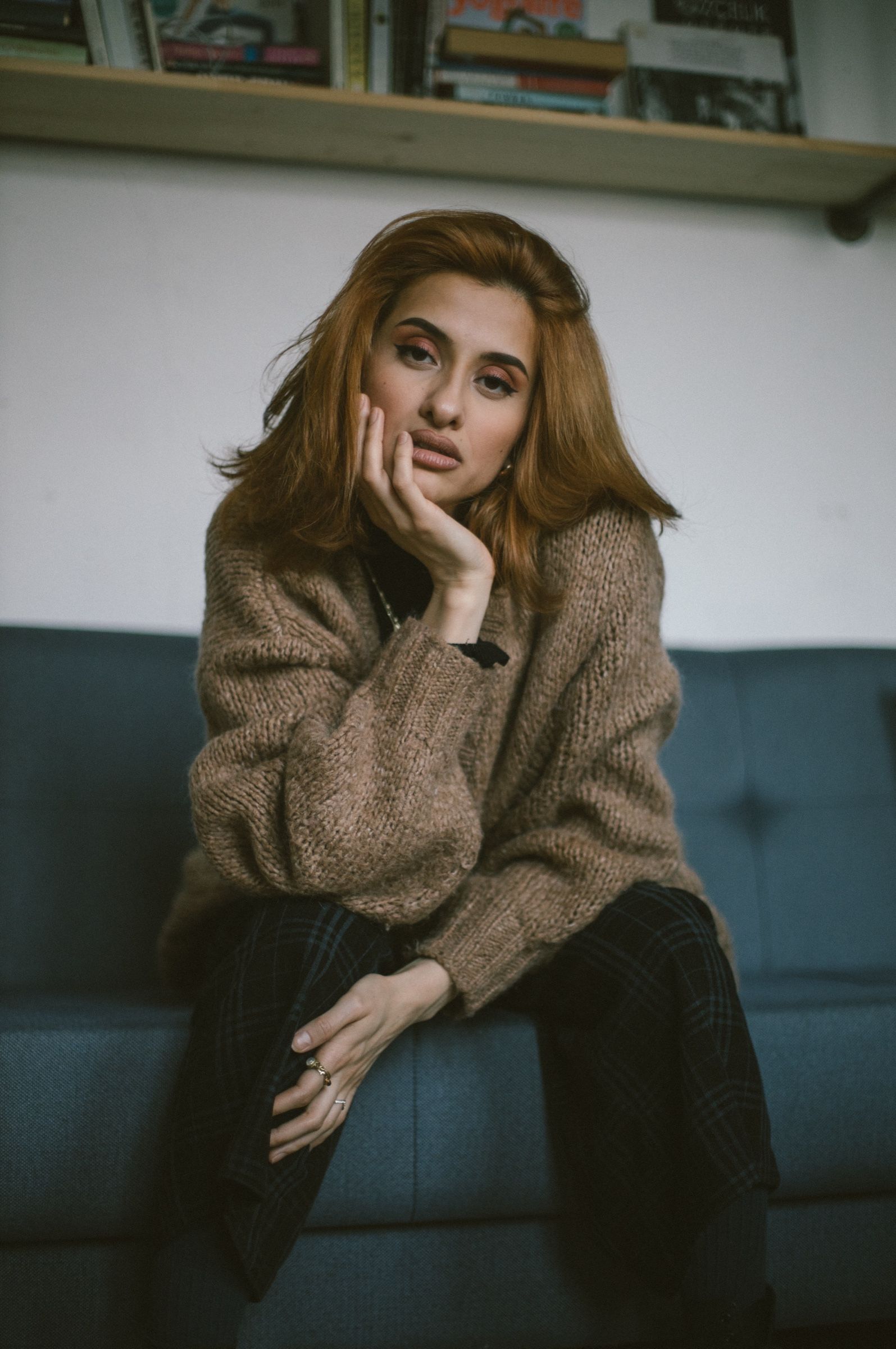 Elif sits on the blue sofa in a wool cardigan and checked trousers, chin resting on one hand, looking straight at the camera.