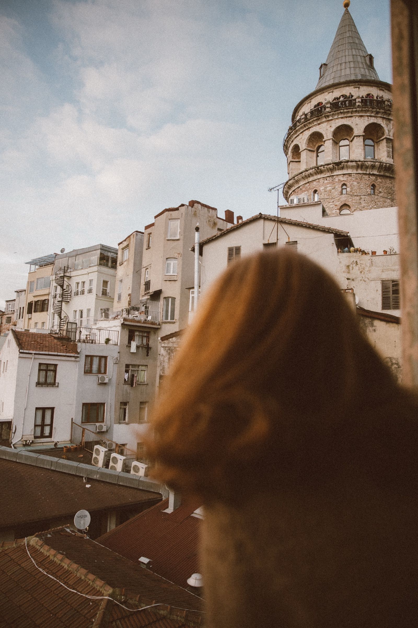 Elif's hair fills the blurred foreground as she faces away, apartment blocks, tiled roofs and the Galata Tower sharp behind.