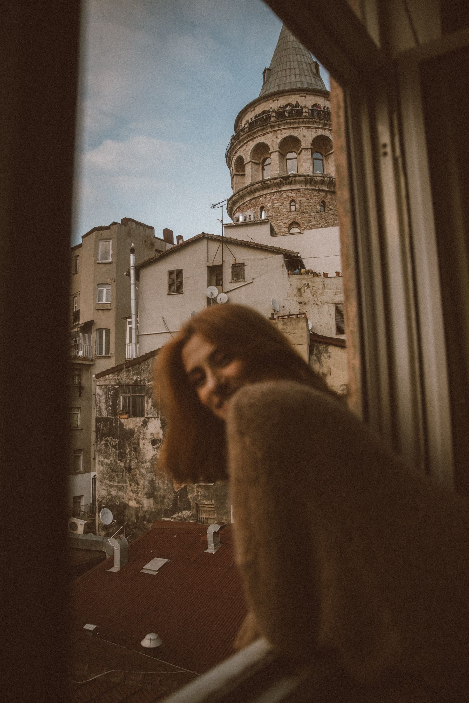 Elif leans out of a tall window with her head tipped and eyes closed, the rooftops and the Galata Tower rising behind her.
