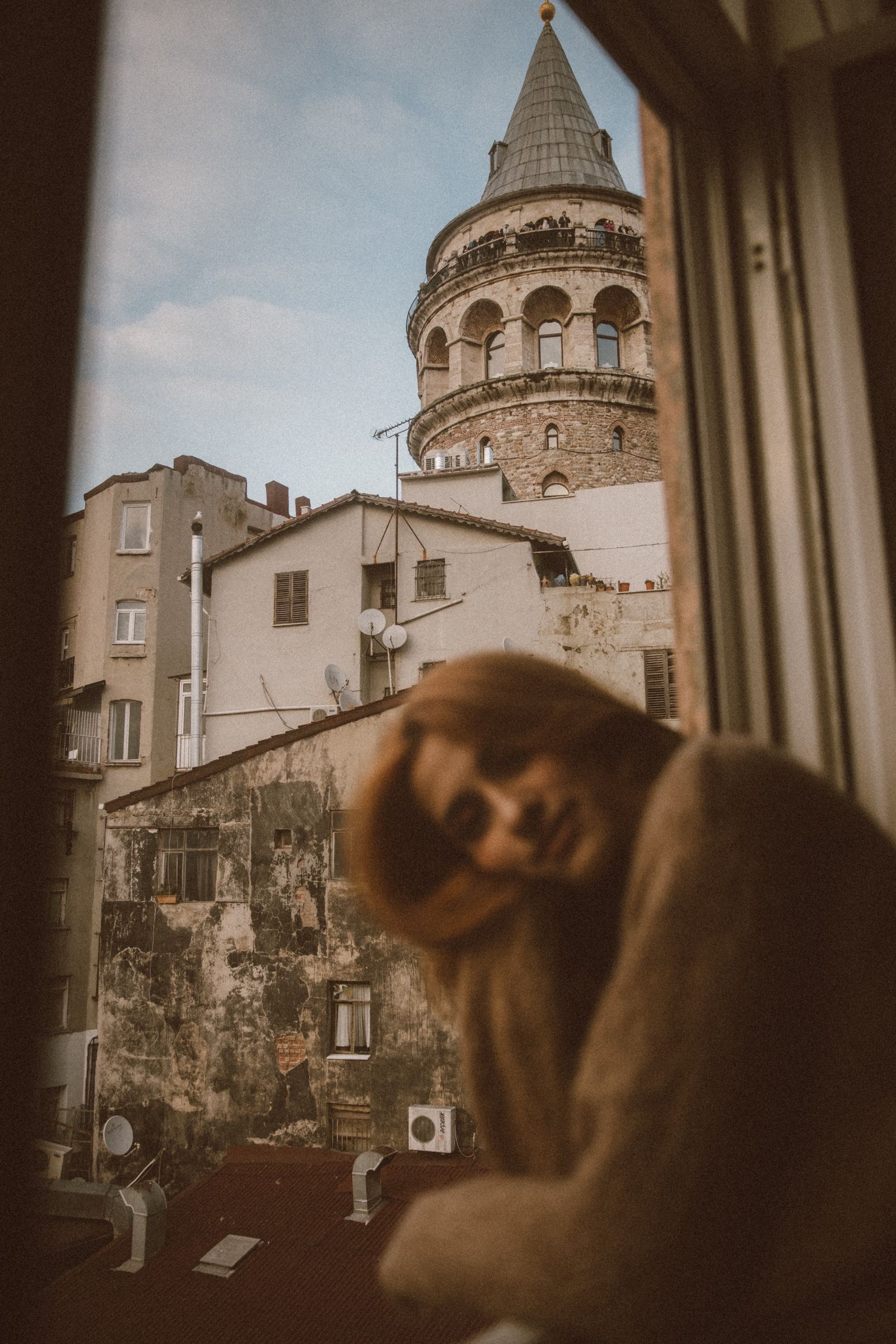 Elif bends low in the blurred foreground with a hand to her face, the Galata Tower sharp above the rooftops behind.