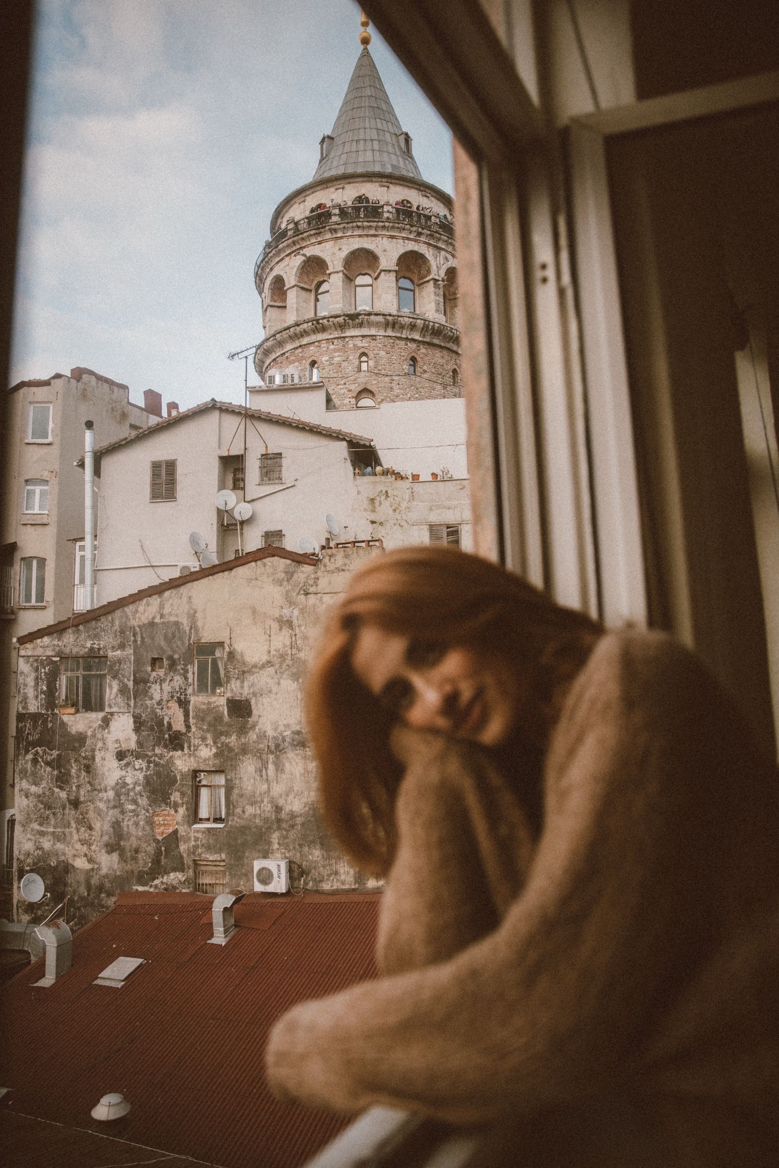 Elif rests her head on her hand at the window, soft in the foreground, with tiled roofs and the Galata Tower sharp behind her.