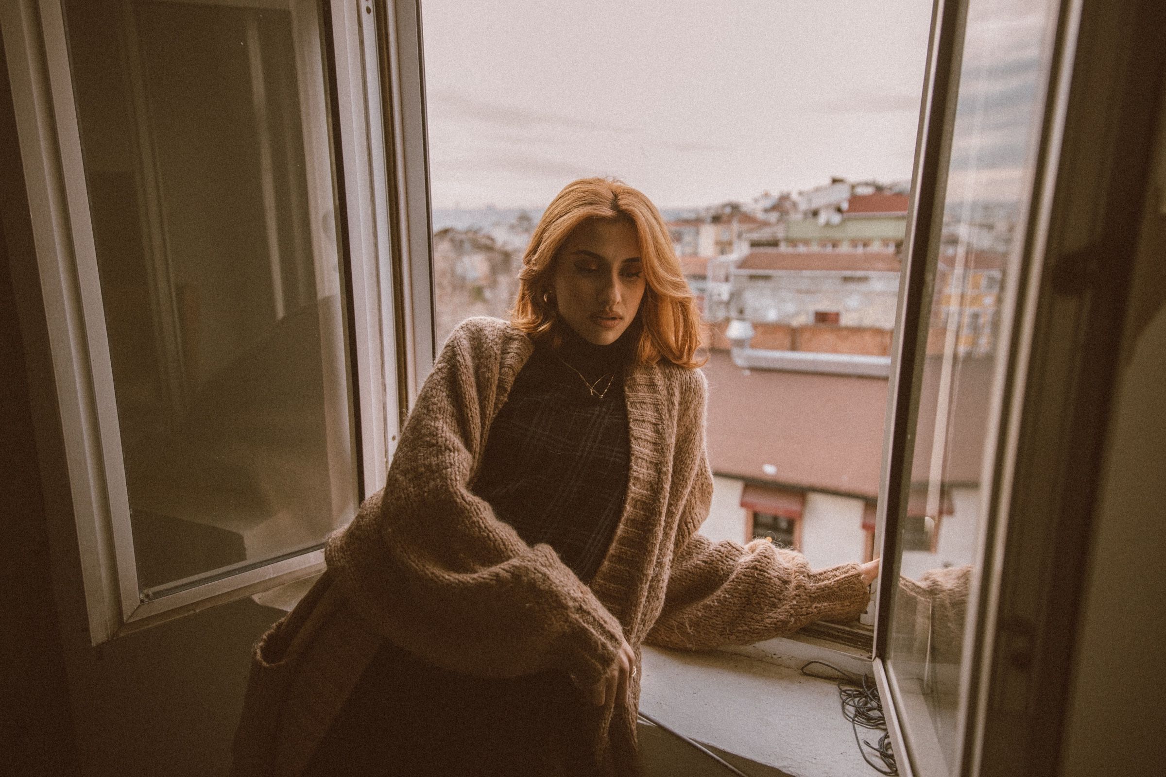 Elif sits in the open window in a chunky knit cardigan, one hand on the frame, looking down, the city rooftops behind her.