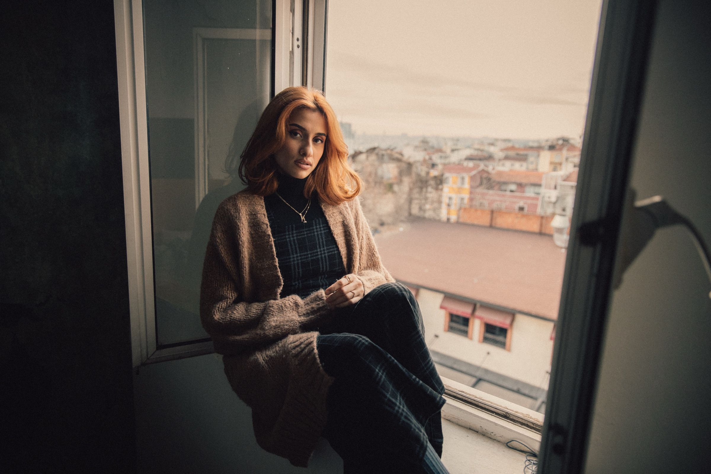 Elif sits in an open window in a knitted cardigan and plaid dress, looking at the camera, rooftops behind her.