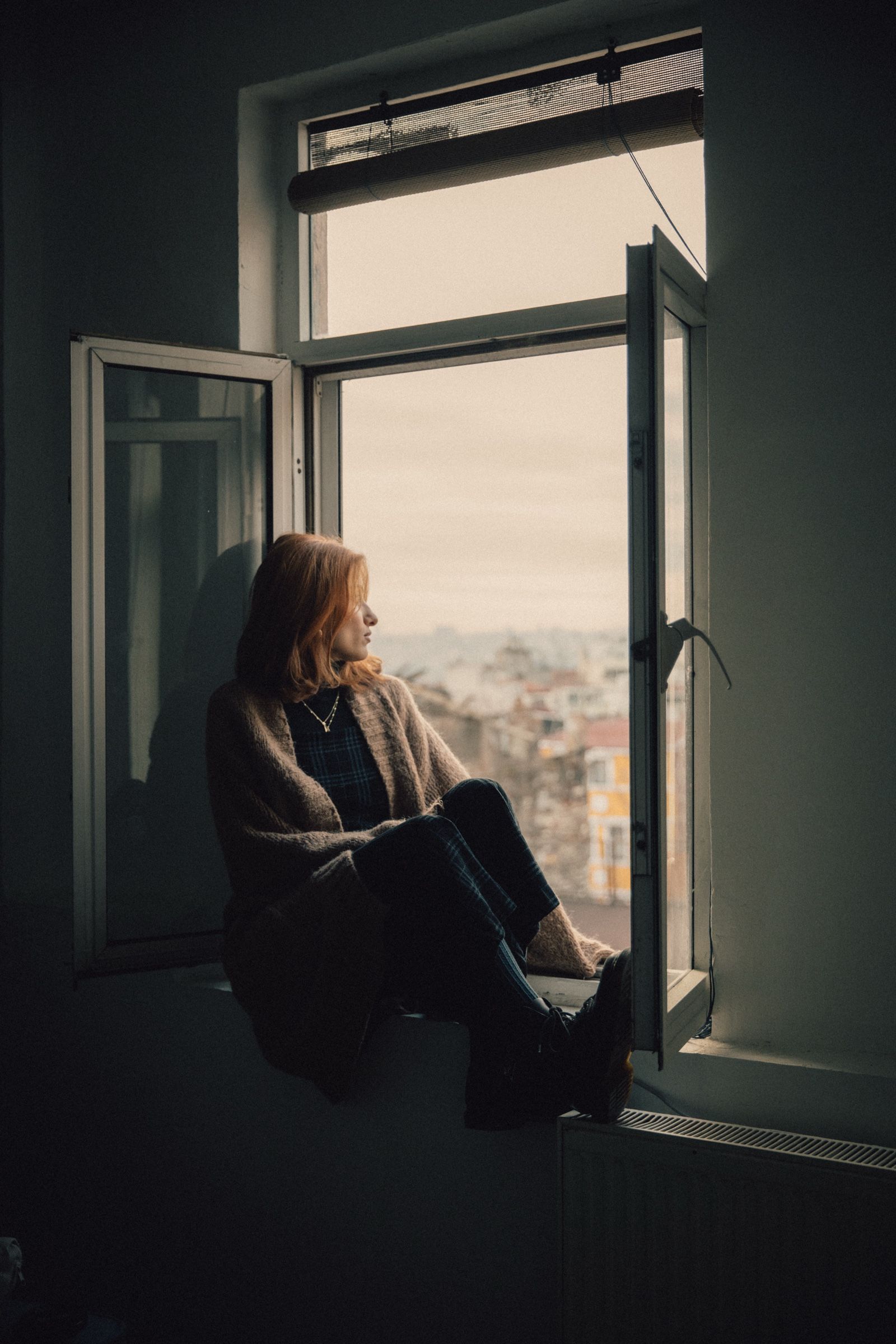 Elif sits on a windowsill in a dim room, knees drawn up, turned toward the daylight and the rooftops outside.