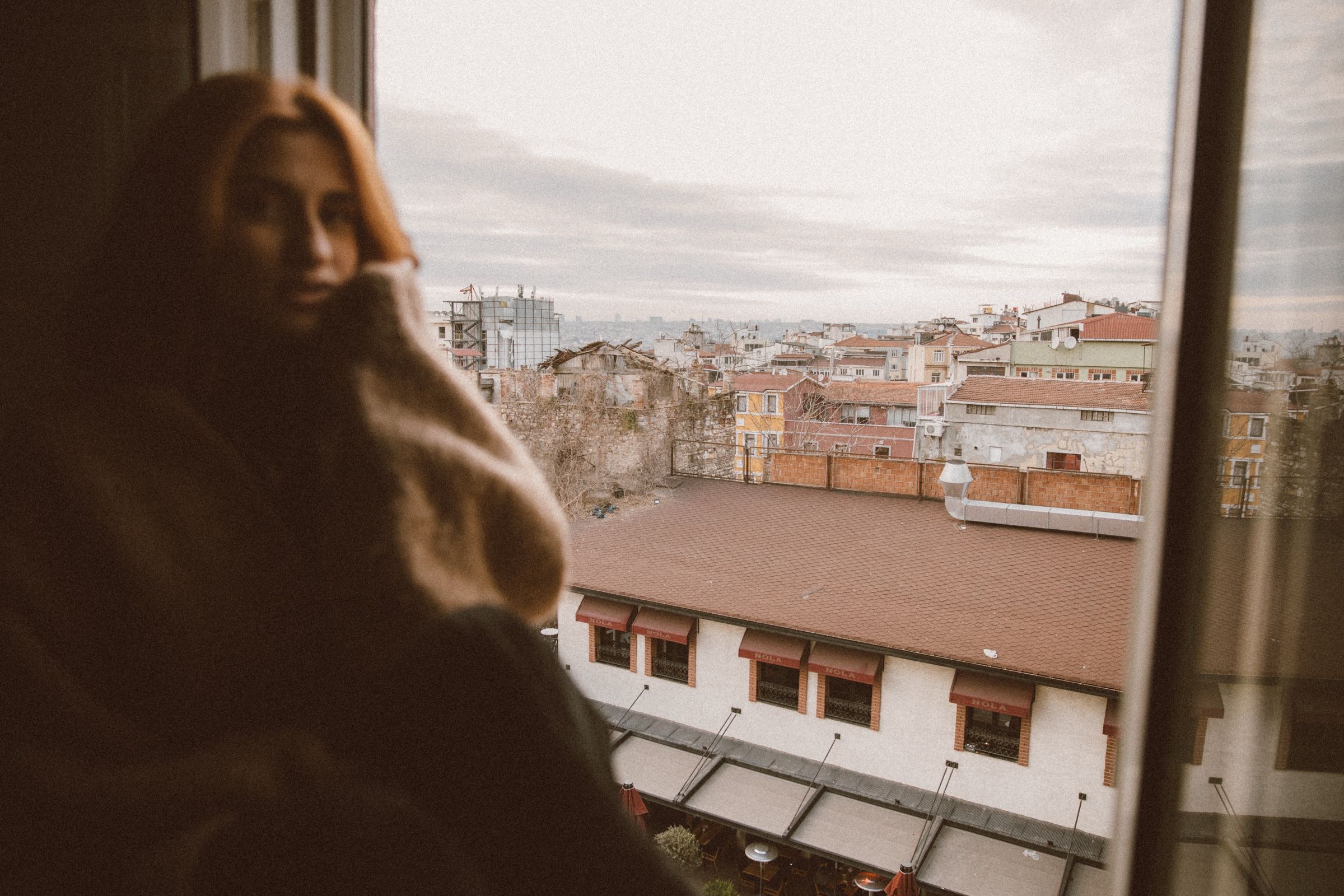 Elif stands close to the glass, blurred and looking back at the camera, the rooftops and hazy skyline sharp beyond.