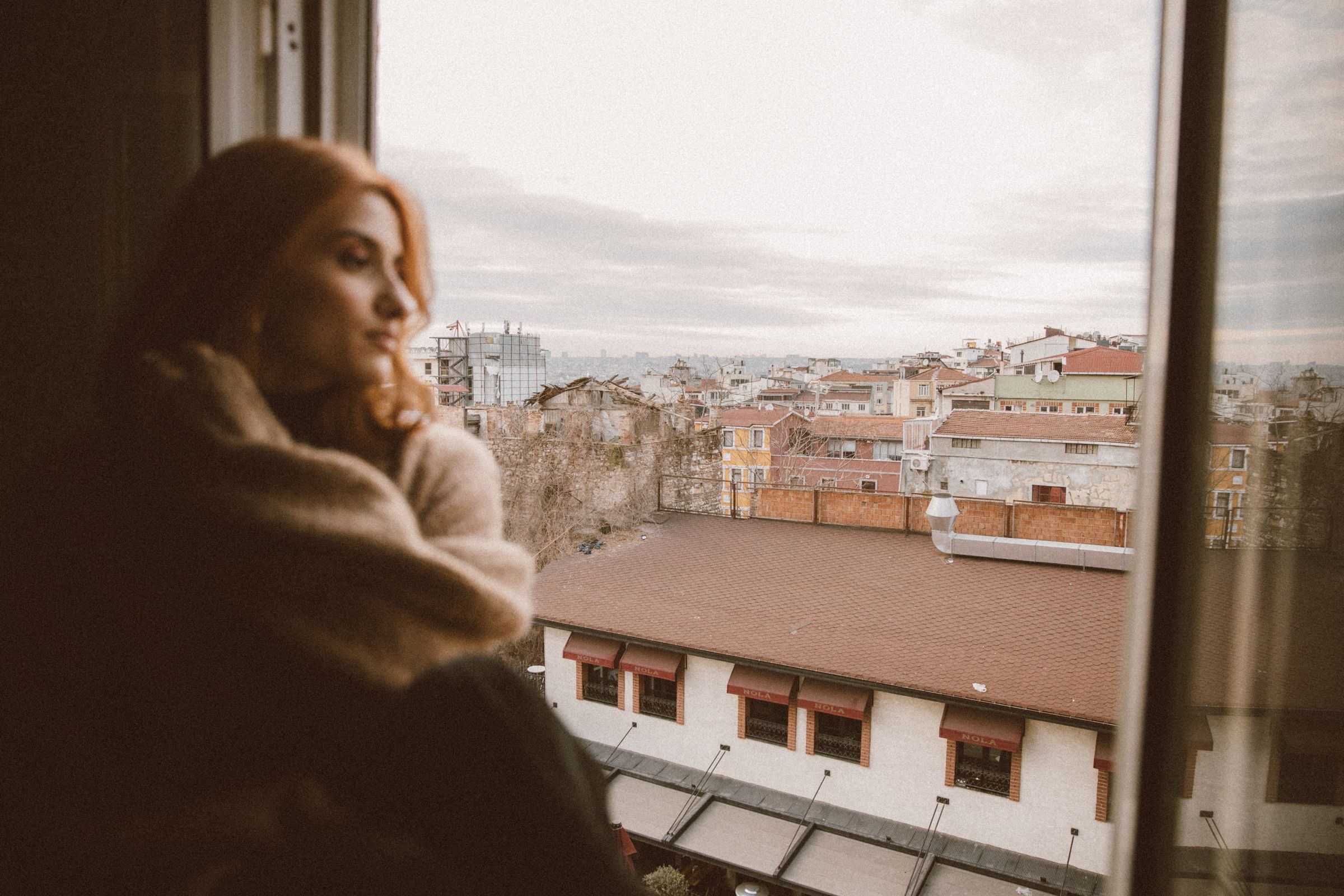 Elif stands at the window in soft focus with her eyes lowered and a hand at her neck, rooftops sharp beyond.