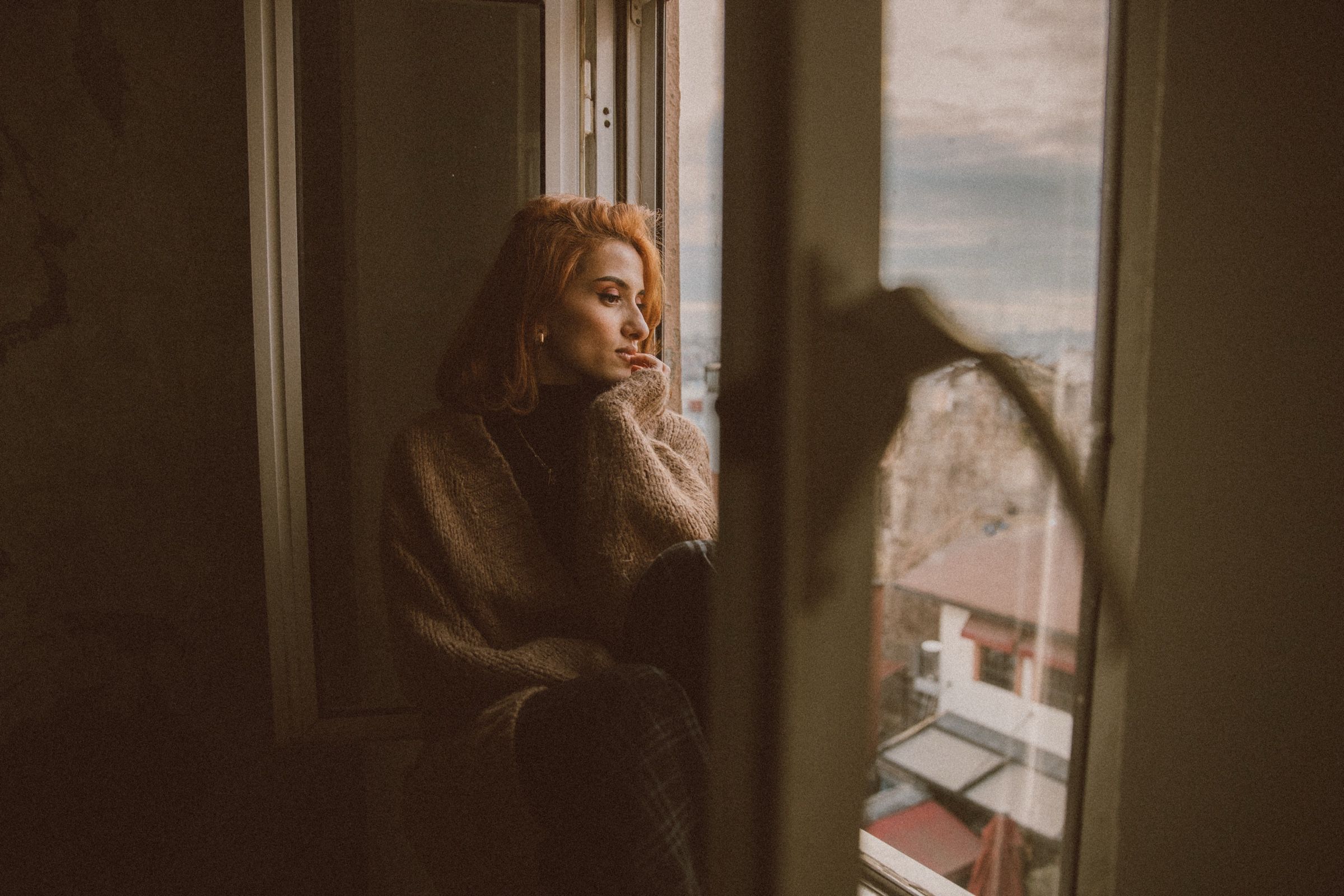 Elif sits by the window in a heavy knitted sweater, hand at her chin, looking out over the rooftops.