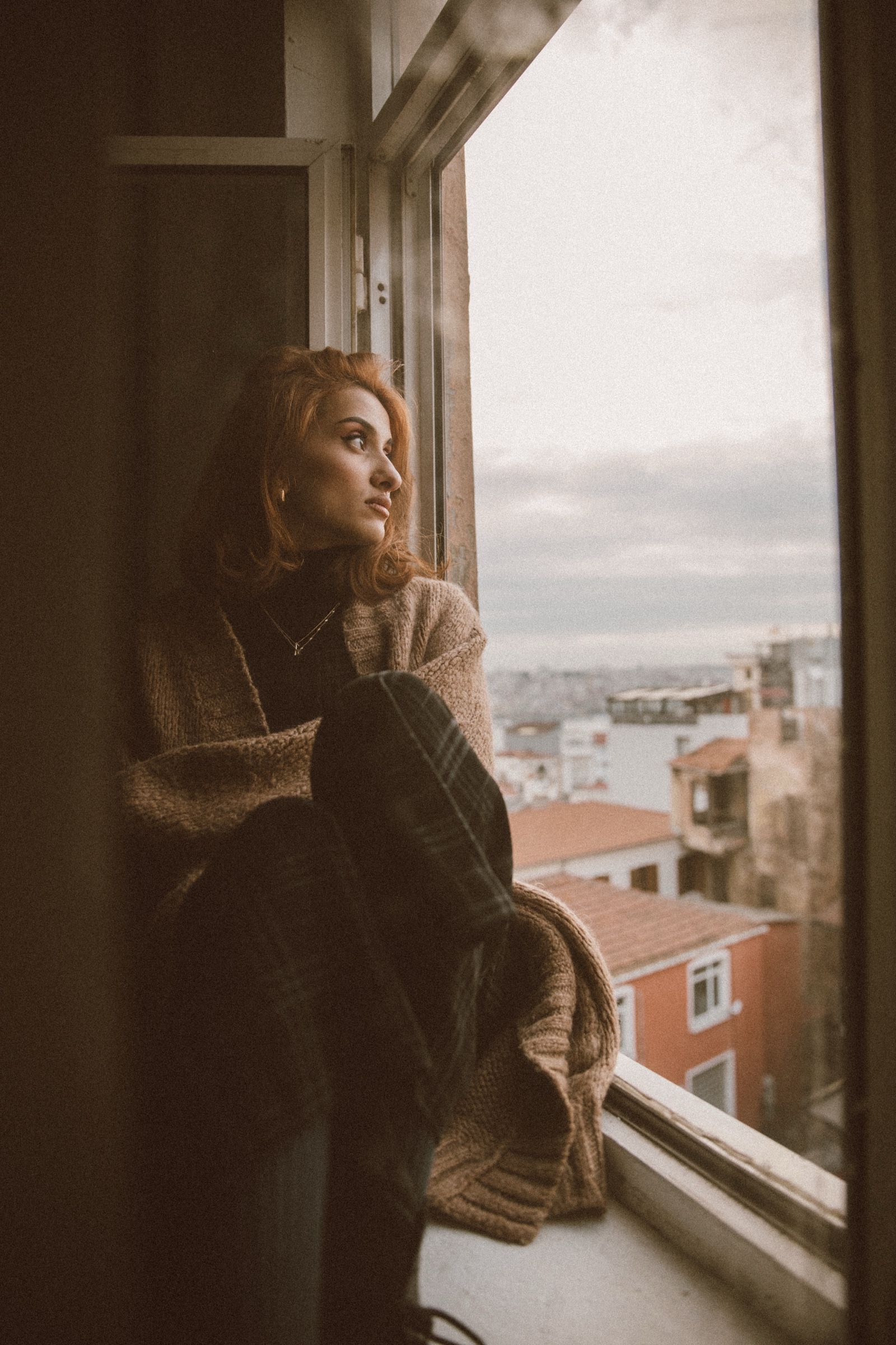 Elif sits in the window recess with her knees pulled up in a knitted sweater, watching the rooftops and pale sky.