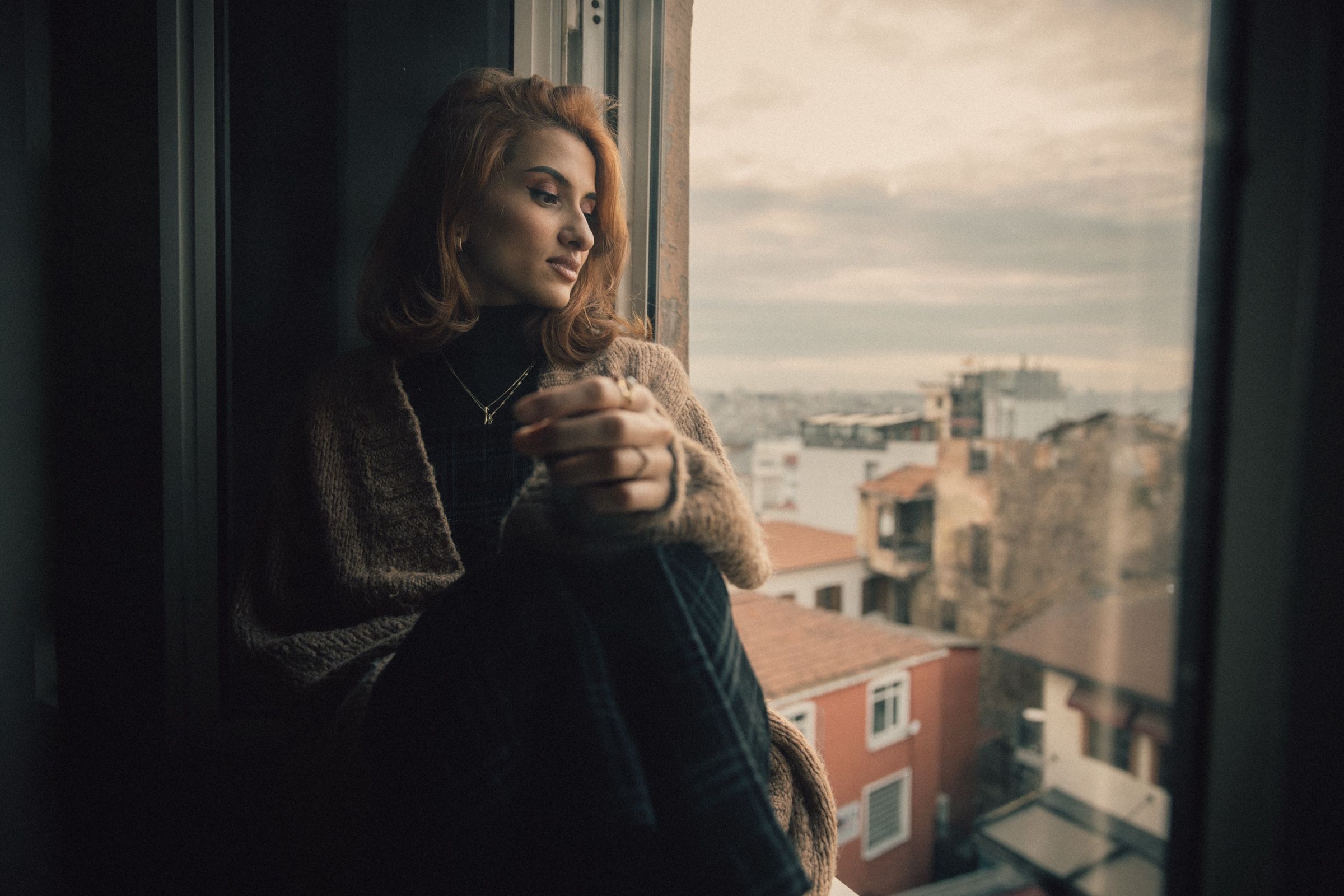 Elif sits at the open window with her arms around one knee, in a black turtleneck and knitted cardigan, the city behind her.