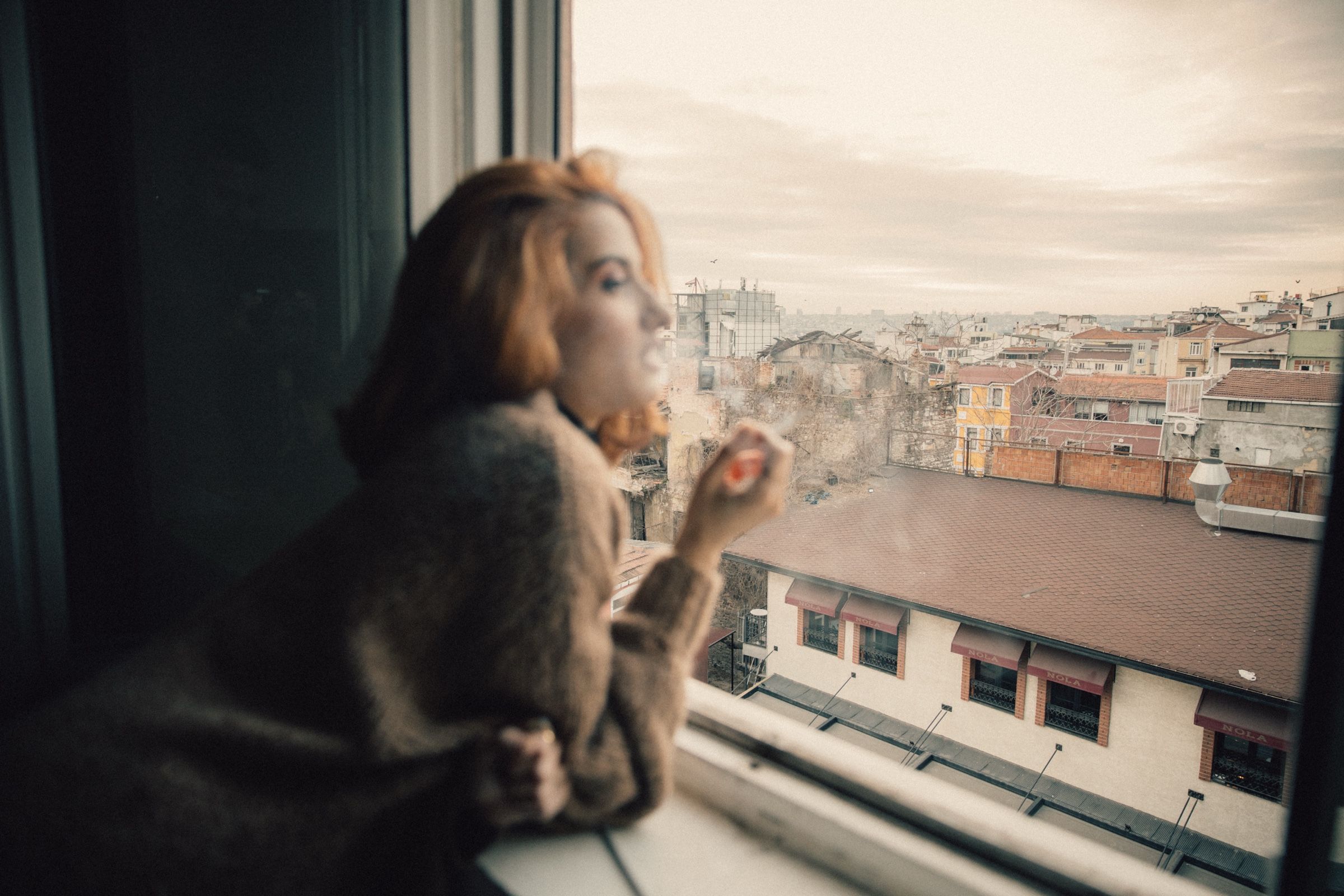 Elif exhales smoke in profile at the open window, out of focus, the rooftops and low buildings sharp below her.