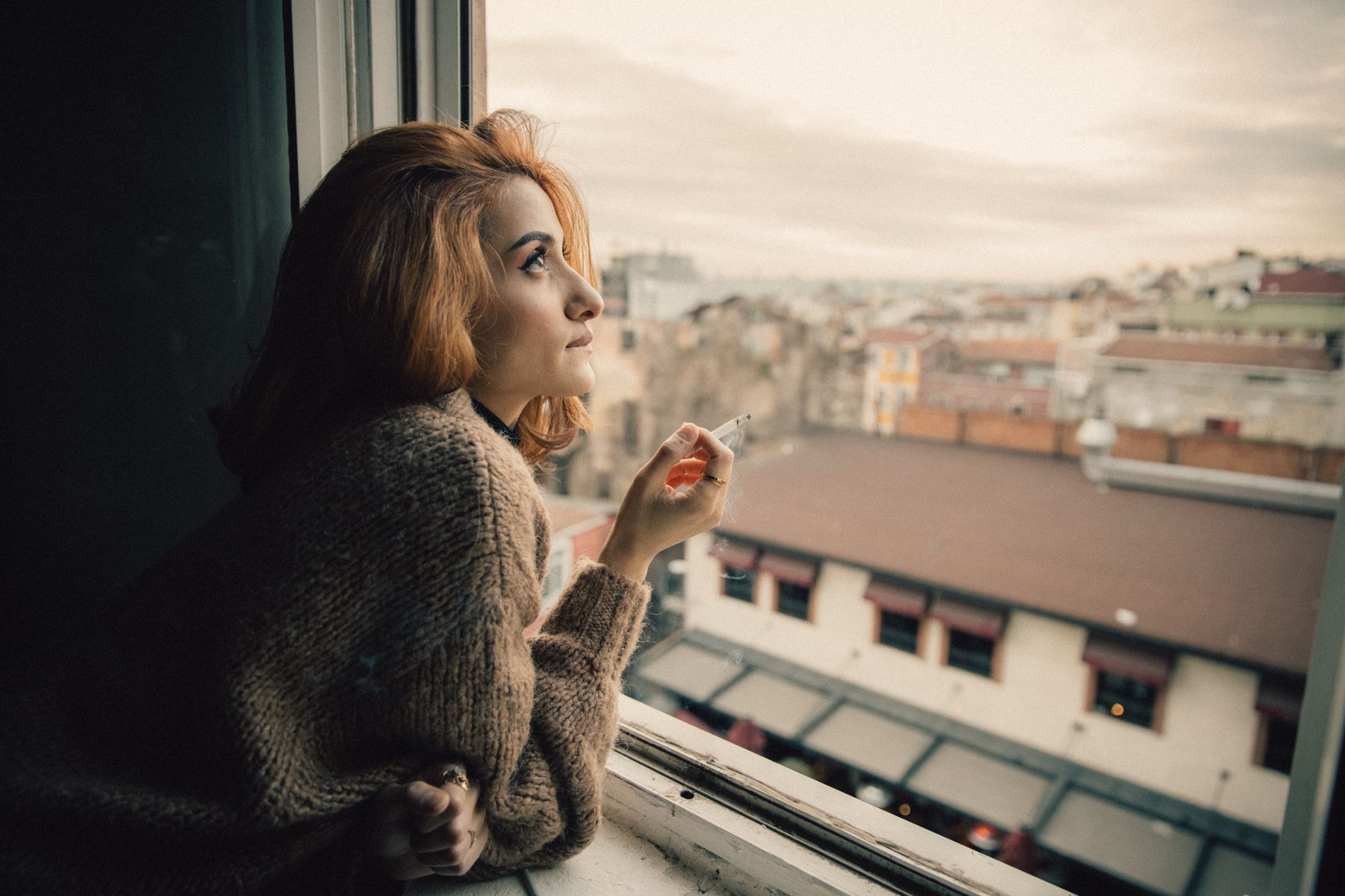 Elif rests her arms on the windowsill in a knitted cardigan, holding a cigarette and looking out across the rooftops.