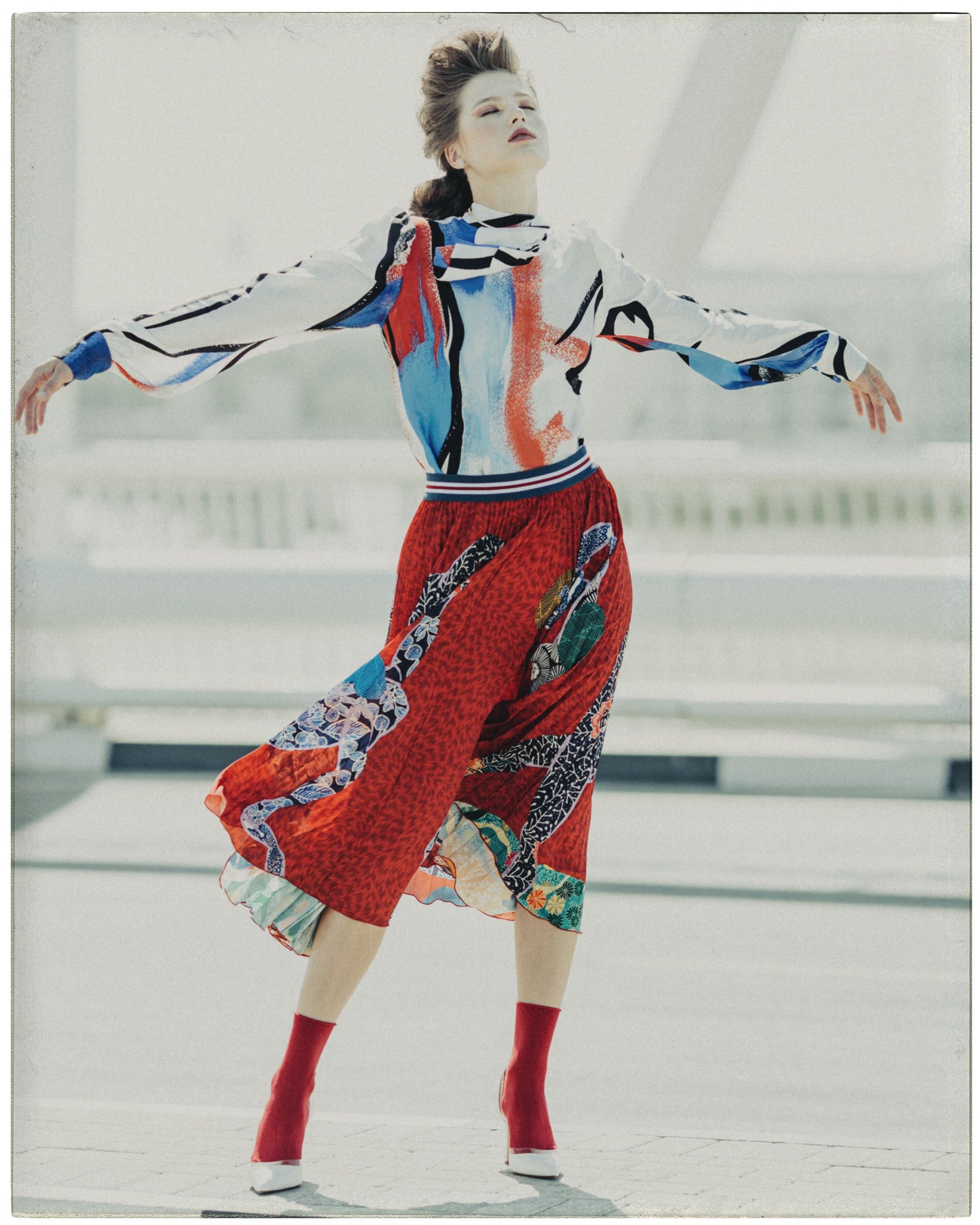 The model spins on a sun-bleached rooftop deck in a white silk blouse printed with red, blue and black brushstrokes, over a red patchwork midi skirt and red sock boots.