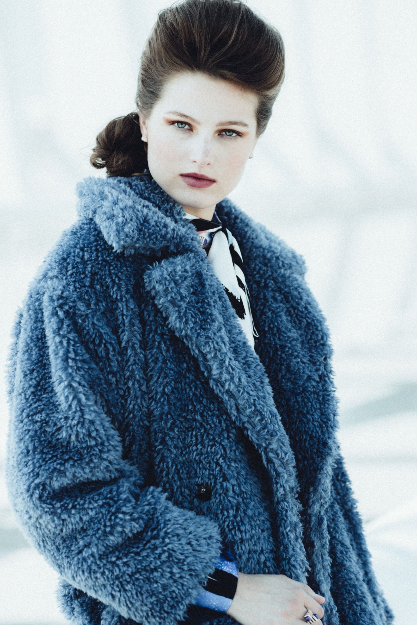 The model turns to camera in an oversized slate-blue shaggy faux fur coat with wide notched lapels, a black and white printed scarf knotted beneath, against a hazed-out sky.