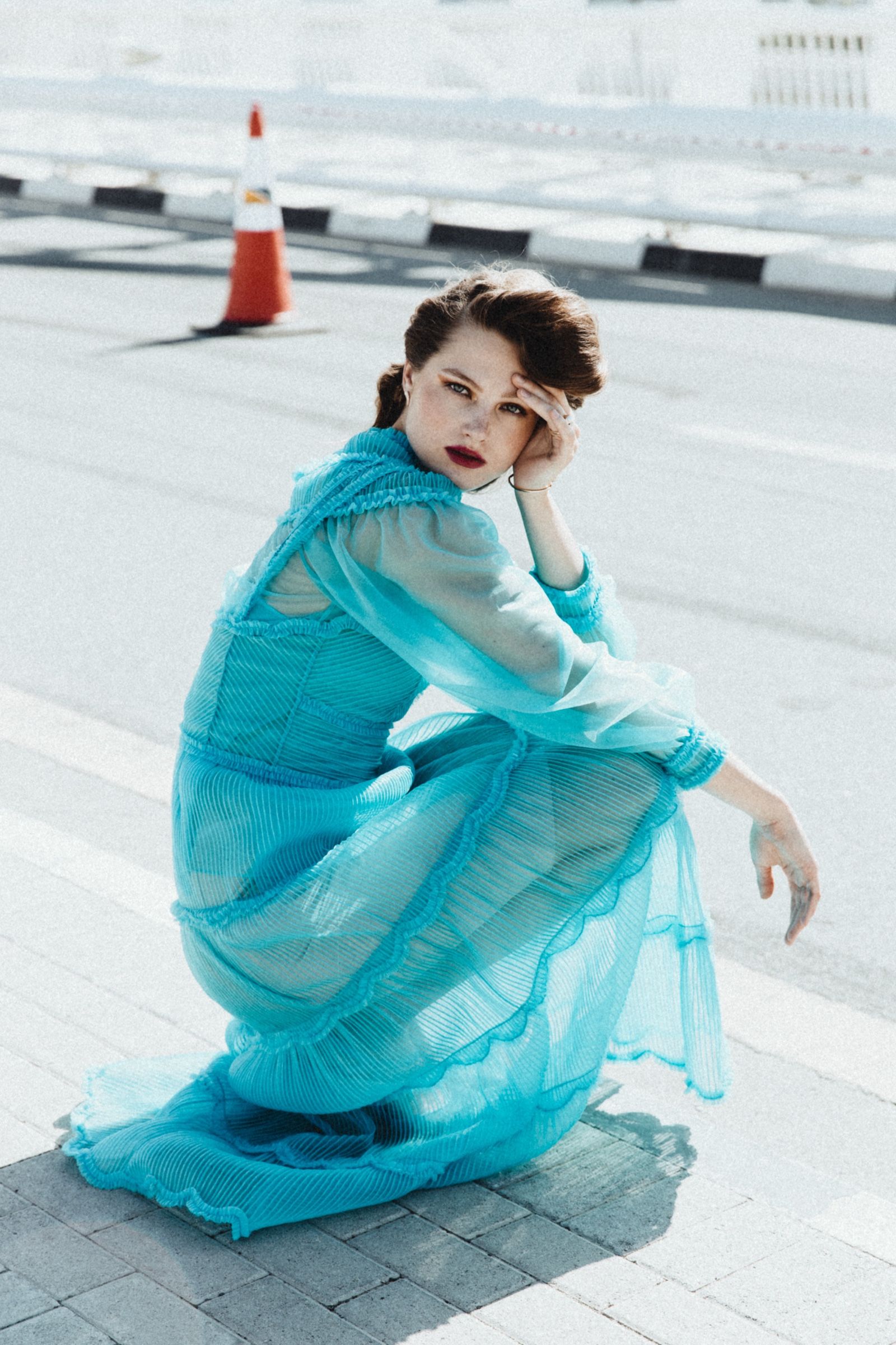 Seated on pale rooftop paving in hard midday sun, she wears the turquoise pleated organza gown, its scalloped tiers and sheer sleeves pooling across the concrete.