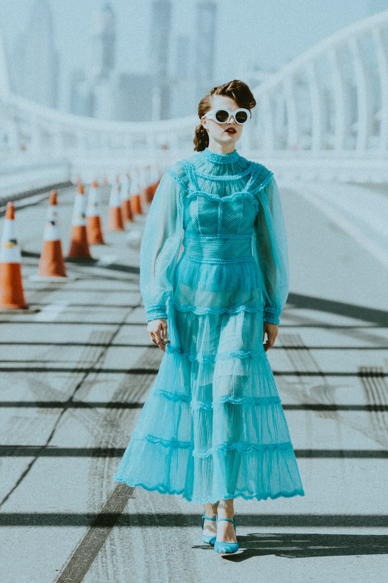 The model walks a rooftop ramp lined with traffic cones in a turquoise tiered organza dress with high ruffled neck and blouson sleeves, white sunglasses, skyline hazed behind.