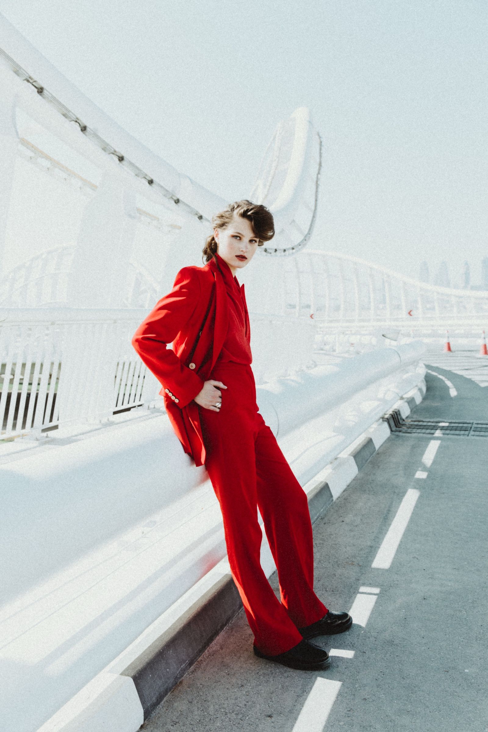 The model leans against a white parapet in a red wool trouser suit and black loafers, flat daylight washing the rooftop road behind her.