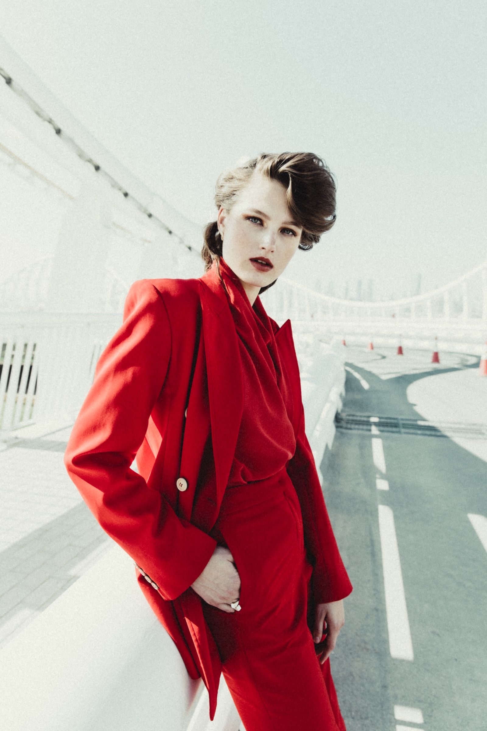 The model stands hand on hip in an oversized red blazer over a draped red blouse, hazy skyline and traffic cones behind.