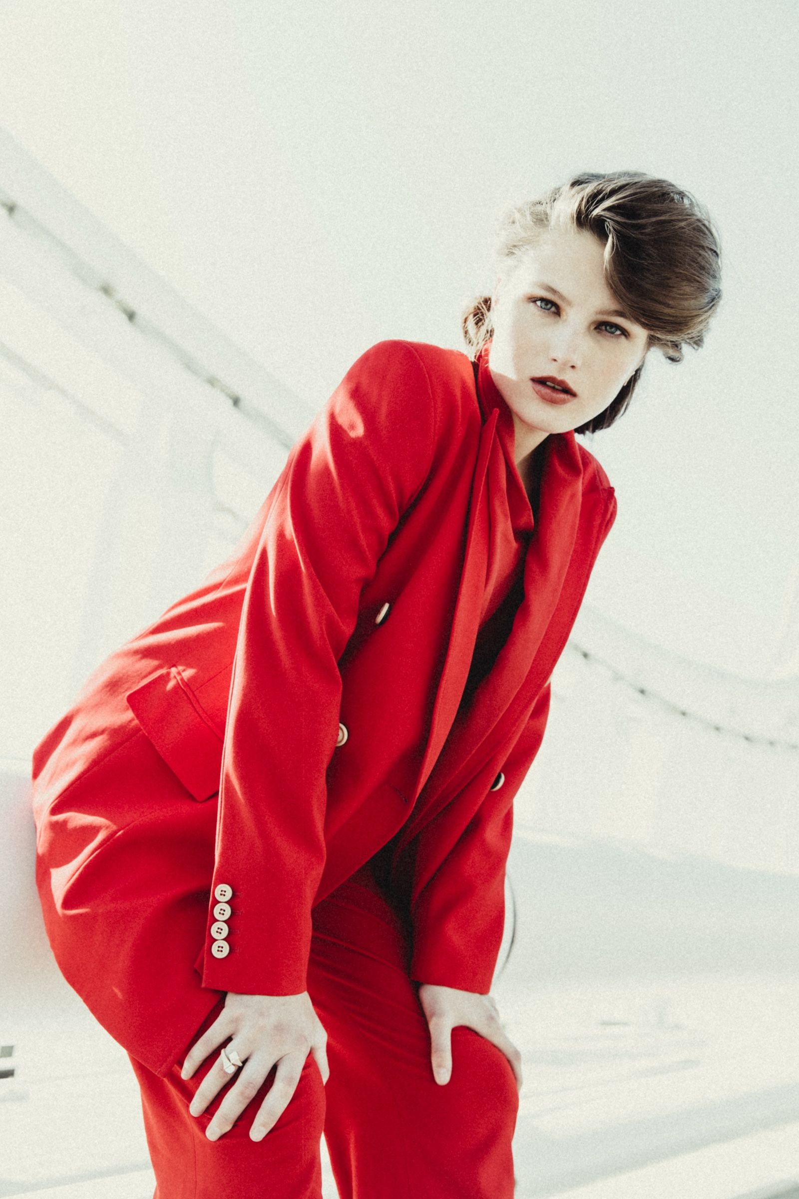 The model bends forward with hands on her knees in a double-breasted red suit, framed against a blown-out white rooftop wall.