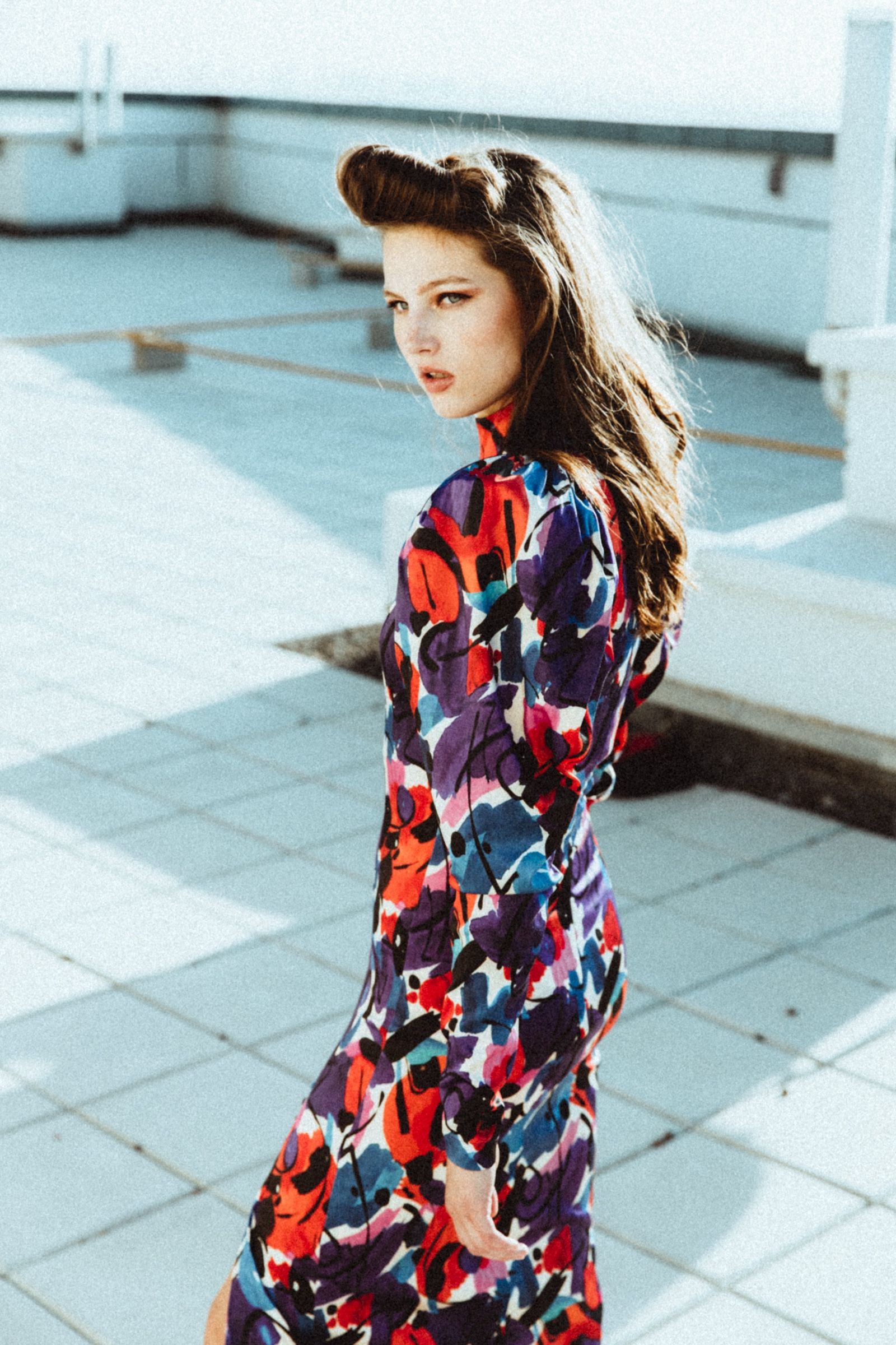 Turning back over her shoulder on pale rooftop tiling, she wears a long-sleeved high-neck dress in red, purple and blue abstract print.