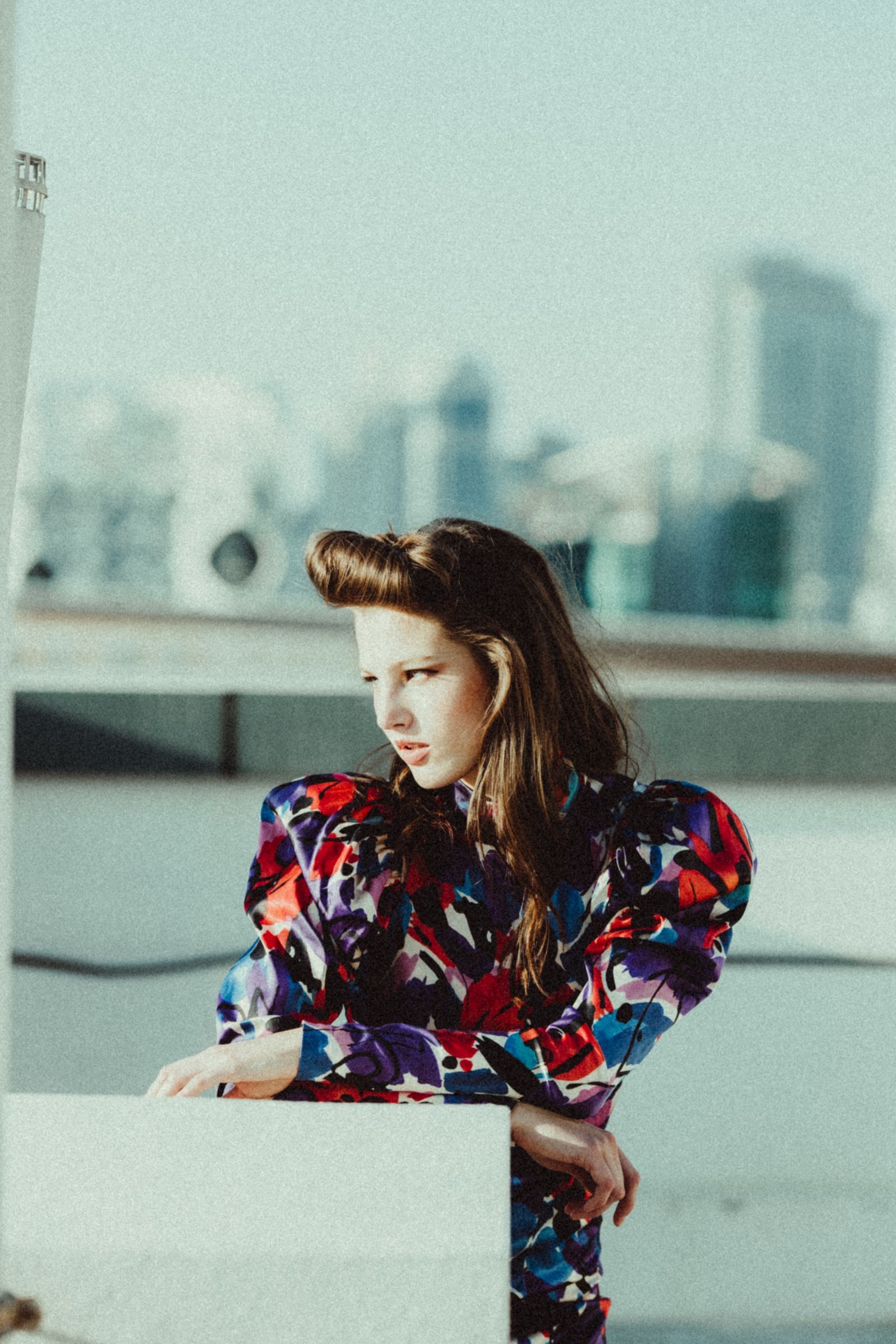The model leans on a low white parapet in a high-necked dress printed with red, purple and blue brushstrokes, hazy skyline behind.