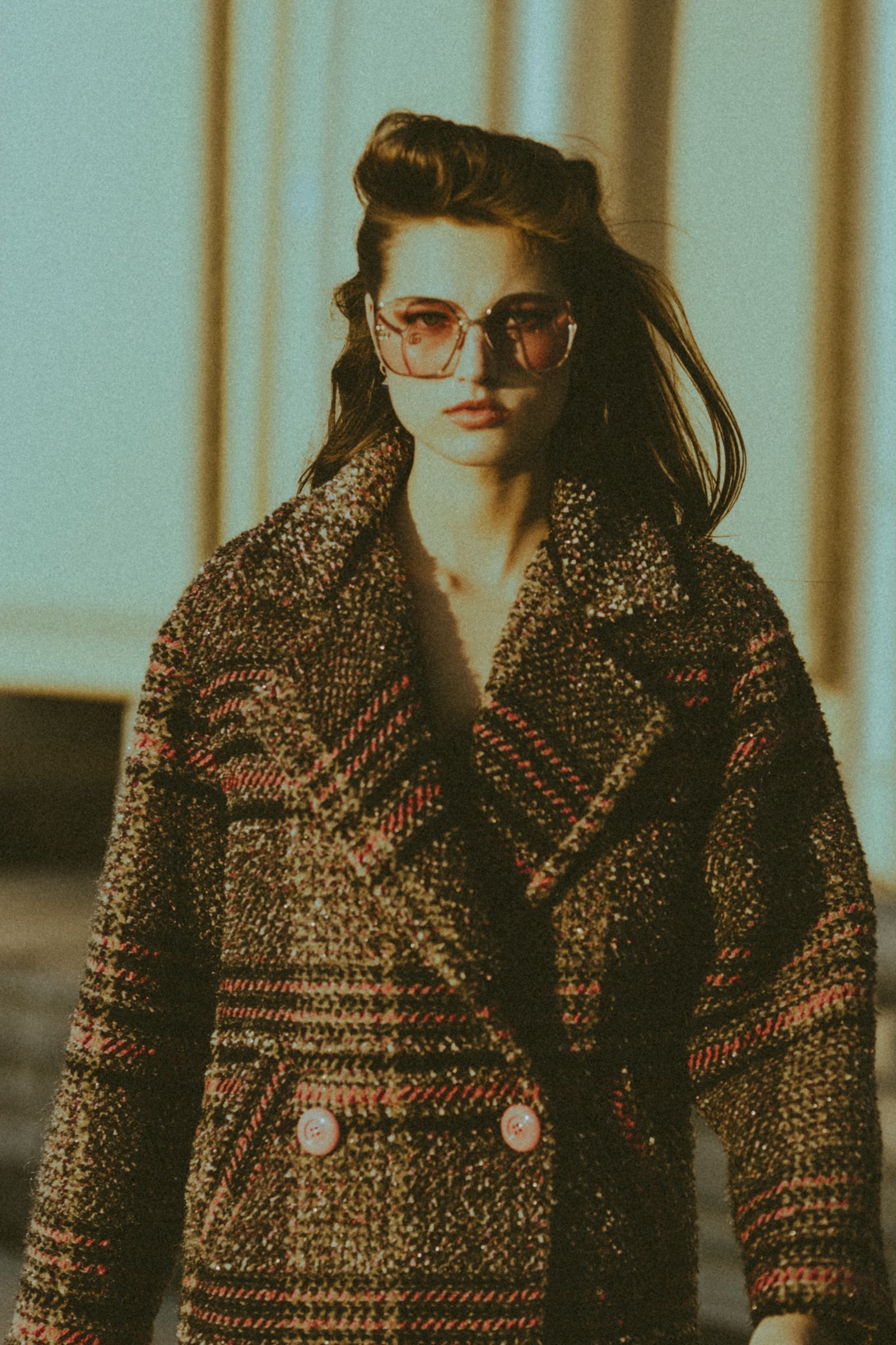 Close waist-up frame of the model in a double-breasted boucle tweed coat with pink checks and oversized buttons, wearing rose-tinted sunglasses in warm low sun.