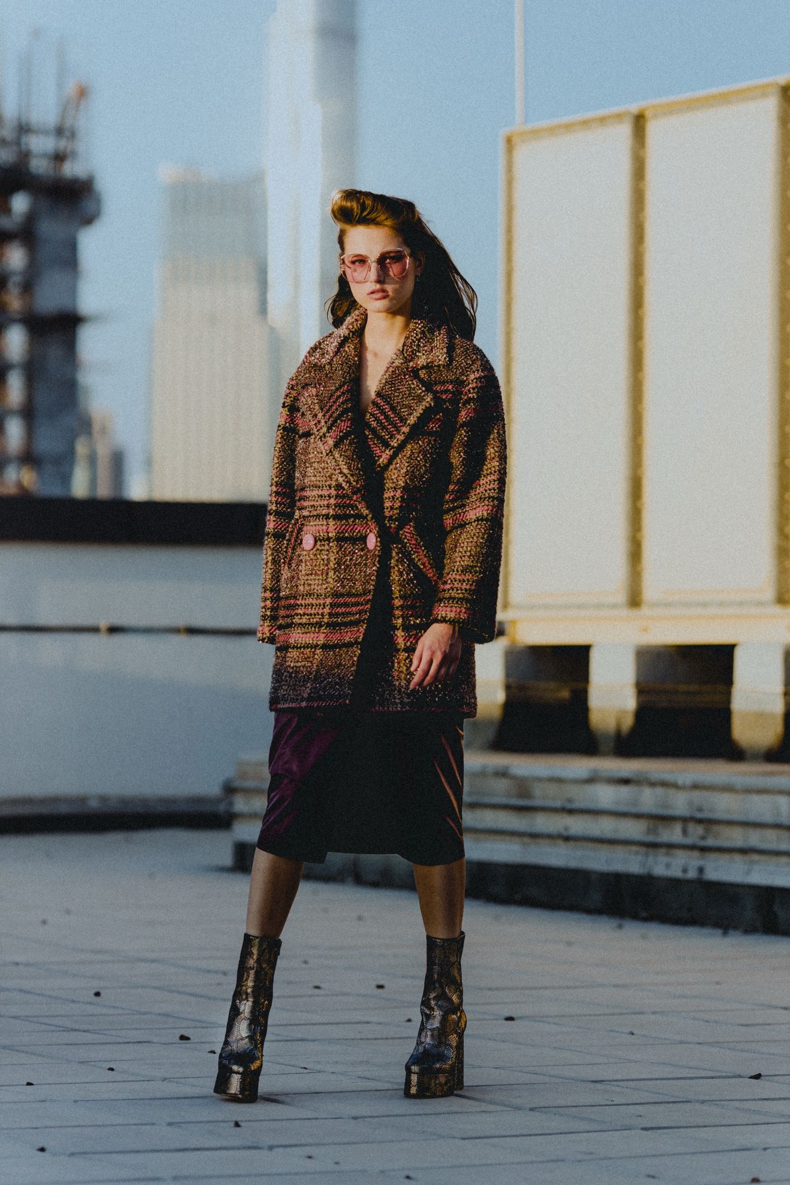 Full-length on the rooftop at golden hour: pink-checked tweed coat over a dark plum skirt, snakeskin platform boots, towers rising behind.